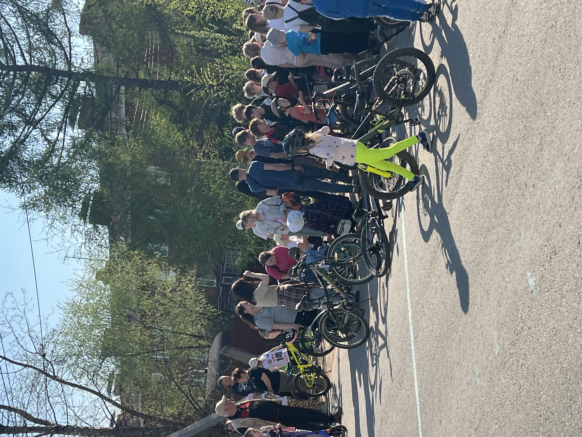 The image depicts a group of people and bicycles on a paved surface, likely a street or a parking area. The location is Noviy, Russia. The group consists of adults and children, many of whom are standing next to bicycles. Some of the bicycles are lined up in a row. The people are casually dressed. The background includes trees, a brick building, and a clear blue sky. The lighting indicates it is daytime.