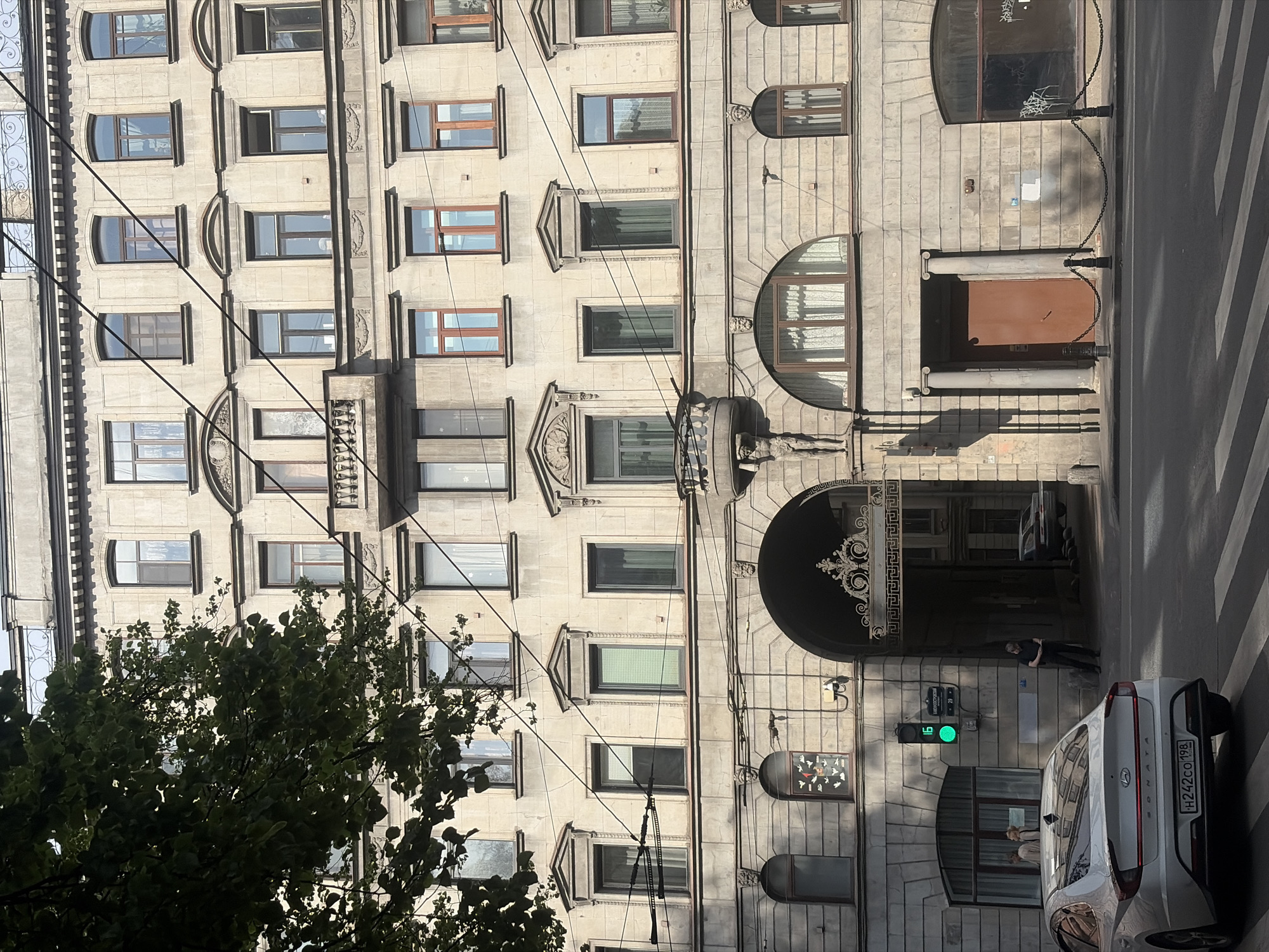 This image depicts the facade of a multi-story historic building located at Nevsky Prospekt, 29, in Saint Petersburg, Russia. The building features a light-colored, ornate stone or stucco exterior with classical architectural elements.
The facade displays numerous rectangular and arched windows across multiple floors. Several windows on upper floors are adorned with decorative pediments or surrounds. A prominent wrought-iron balcony with a balustrade is situated on the third floor from the top. A smaller, ornate balcony on the second floor from the bottom is supported by a sculpted male figure (atlante).
At ground level, a large, dark arched gateway leads into a passage or courtyard, featuring decorative iron gates. To the left of the archway, a large semicircular window with white curtains is visible. Above this window, a smaller window displays an art installation composed of multiple white bird-like cutouts on a dark background. A green traffic light is affixed to the wall next to the archway, displaying the number "16" in green. To the right of the archway, a brown wooden door is flanked by two columns, with black bollards connected by chains guarding its front. Further to the right, another arched window appears boarded up or dark, showing some white graffiti.
In the foreground, a white Hyundai Sonata sedan (Russian license plate H242CO 198) is positioned on a paved street with a white zebra crossing. The car is facing away from the viewer, towards the right.
Two individuals are partially visible inside the semicircular window to the left of the archway. A man in dark clothing stands within the archway's entrance, looking down at an object in his hands. A dark-colored car is parked inside the archway's passage.
A large tree with green foliage occupies the upper left portion of the frame, casting shadows. Multiple black overhead wires diagonally traverse the image across the building's facade. The scene is illuminated by natural light, casting distinct shadows on the building's right side and by the tree.