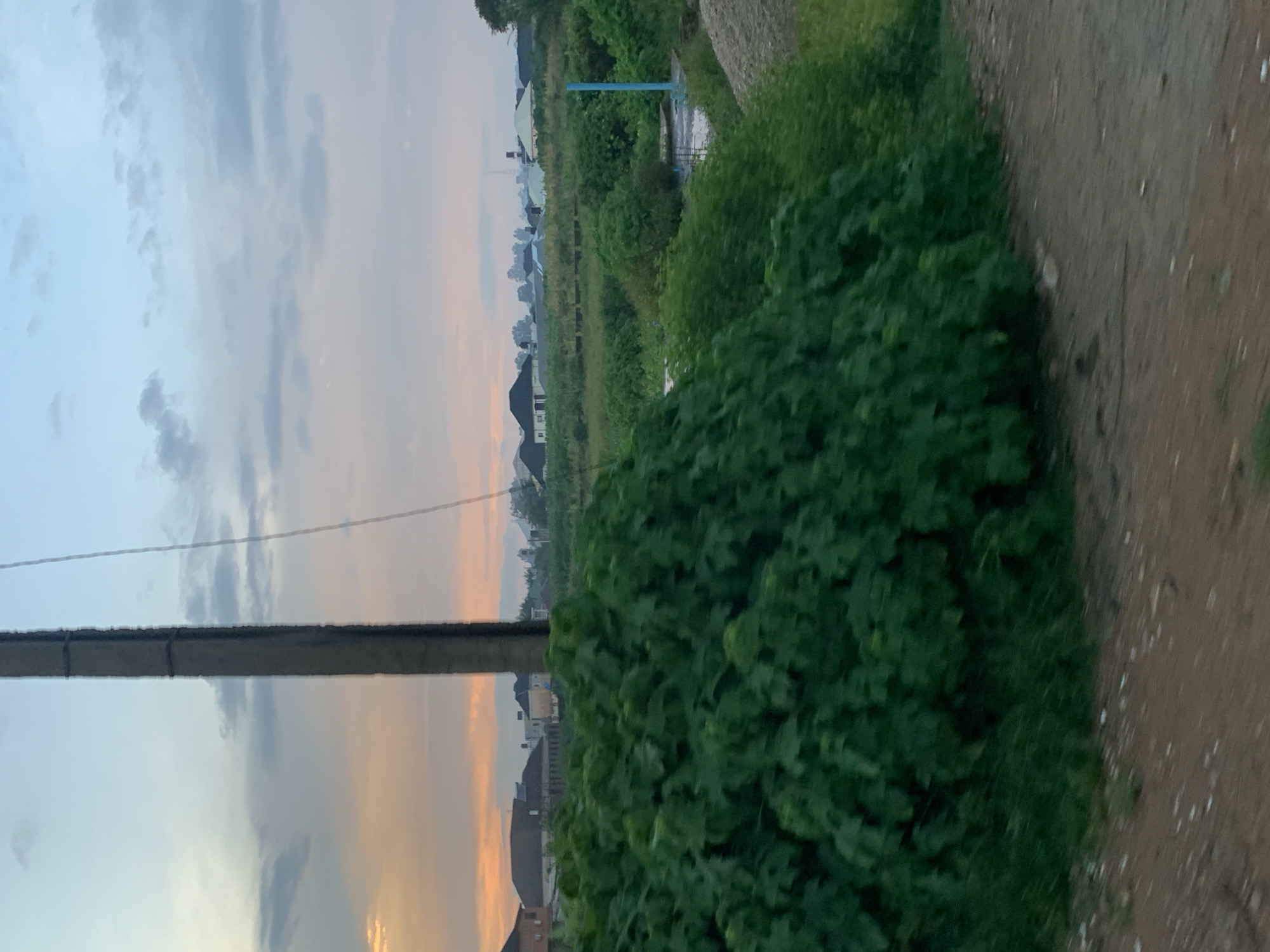 The image depicts a rural landscape at dusk, with a prominent utility pole on the left, bisecting the frame. The sky transitions from a pale blue at the top to hues of orange and pink near the horizon, suggesting the setting sun. In the midground, a dense cluster of green foliage dominates the foreground, obscuring much of the scene. Beyond the foliage, several houses with peaked roofs are visible, interspersed with trees and fields. A dirt road runs along the bottom of the image. The overall impression is of a quiet, undeveloped area, possibly on the outskirts of a settlement. The scene is situated in the city of Gura Top, Nigeria.