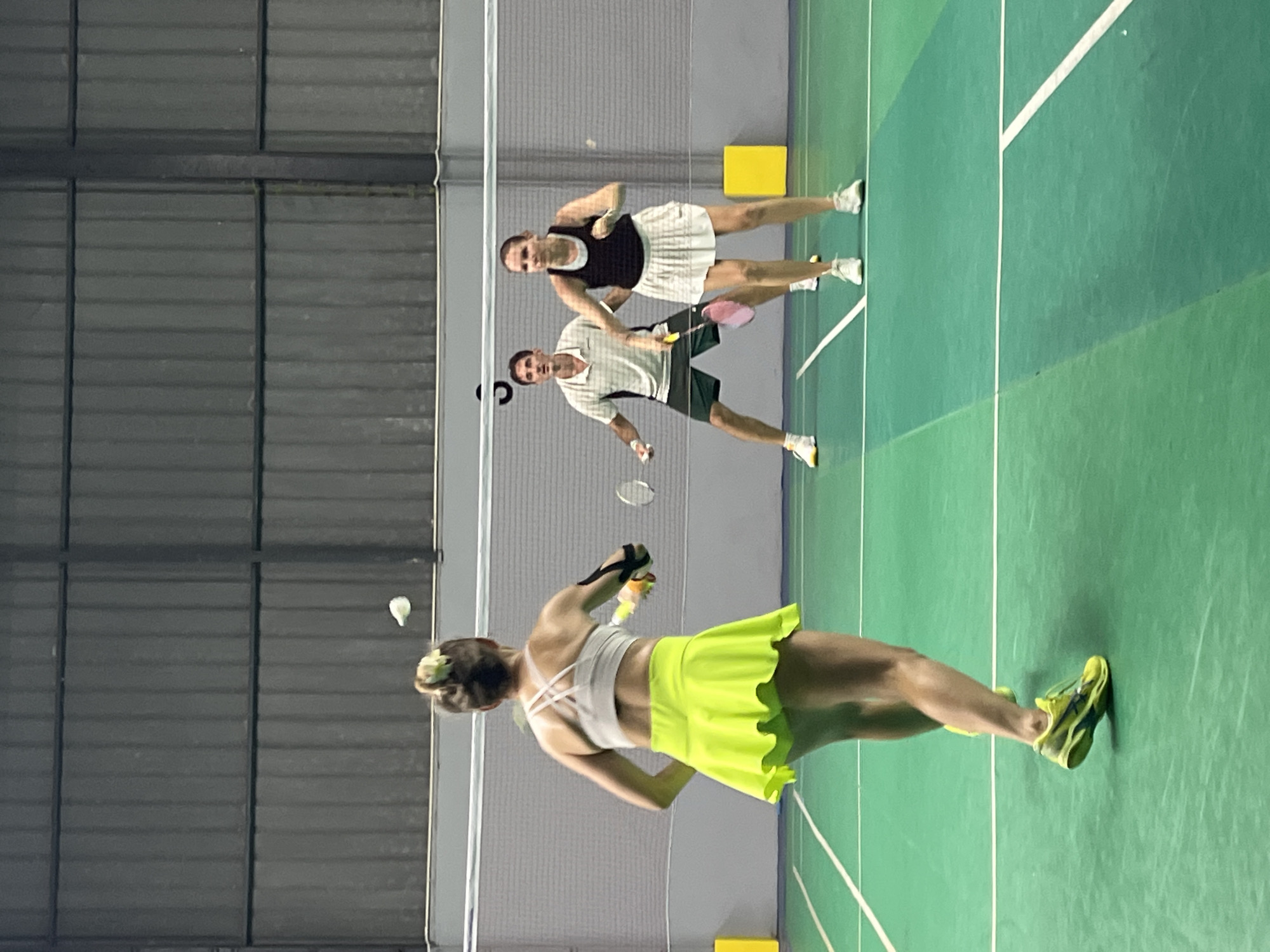 In a badminton court in Ban Rak, Thailand, four individuals are engaged in a doubles match. In the foreground, a woman with her back to the camera, wearing a neon yellow skirt and white sports bra, is poised to hit a shuttlecock. To her right, a man in a striped polo shirt and dark shorts, and a woman in a black tank top and white skirt are also actively playing. A fourth player, partially obscured, is on the far side of the court. The court is marked with white lines on a green surface, and a net divides the playing area. The background features a grey corrugated metal wall.