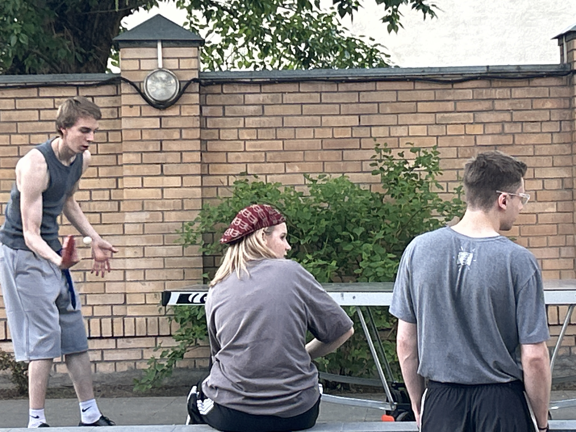 The image shows three individuals outdoors near a brick wall and lush green bushes. A man on the left is dressed in a dark gray tank top and light gray shorts, preparing to hit a table tennis ball with a red paddle. A woman wearing a red bandana sits with her back to the camera, wearing a gray t-shirt and black pants. To the right, a man with glasses is seen from the back, wearing a gray t-shirt and black shorts. A table tennis table is visible behind the woman. A streetlamp is mounted on the brick wall. The location is an urban area in a city of Unknown, Unknown.