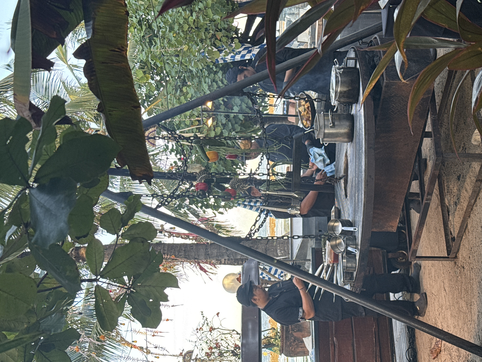 The image depicts an outdoor kitchen setup in Denpasar, Indonesia. Two individuals wearing black uniforms and hats are actively cooking. One individual is focused on the food preparation, while the other appears to be engaged in tasks associated with cooking tools.
Various kitchen utensils, including pots and pans, are visible on a metal structure. Fresh produce, such as vegetables, is also hanging from the structure. The surroundings feature lush tropical plants and trees, including banana leaves and palm trees. In the background, a glimpse of the ocean is visible. The scene suggests an open-air cooking demonstration or a restaurant setting with a focus on outdoor food preparation.