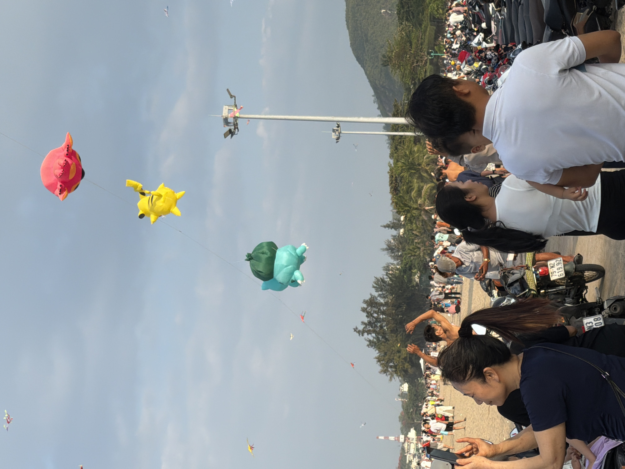 The image depicts a beach scene at sunset in Nha Trang, Vietnam, likely at a public gathering or festival. The sky is overcast with a light blue hue, and the sun is low on the horizon. Several kites are visible in the sky, including a large, pink, octopus-shaped kite, a yellow Pikachu kite, and a teal turtle-shaped kite with a green sack on its back. These kites are strung together on a line, suggesting a kite display or competition.
In the foreground, a woman is taking a photo with her phone, holding it up to capture the scene. She is wearing a dark blue t-shirt. Behind her, a crowd of people is visible on the sandy beach. Many of them are dressed casually in light-colored clothing. There are numerous motorcycles parked in rows, indicating a popular spot or event. The background features palm trees and hills, characteristic of a coastal Vietnamese landscape. Two tall lampposts stand prominently in the mid-ground, one of them adorned with small flags. The overall atmosphere appears lively and communal, with people enjoying the outdoors and the spectacle of the kites.