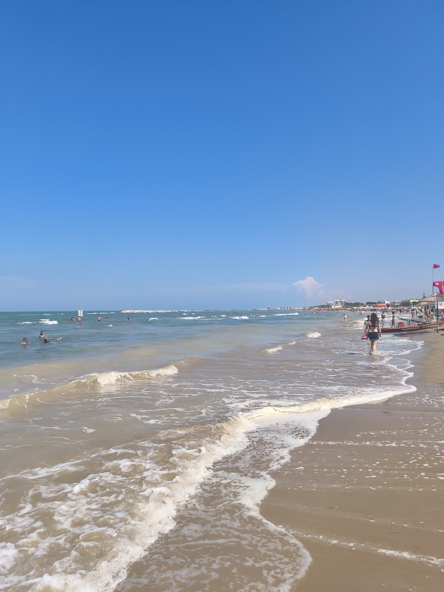 The image depicts a beach scene on a sunny day with a clear blue sky. In the foreground, gentle waves with white foam are washing ashore onto a sandy beach. A woman in a black bikini is walking away from the camera along the water's edge, carrying what appears to be a surfboard or a similar flat object.
Further out in the water, several people are swimming and enjoying the sea. In the distance, a long line of beach umbrellas and buildings suggests a developed coastal resort area. A red flag, possibly indicating a lifeguard station or a warning, is visible on the right side of the image, attached to a structure with the word "SALVATAGGIO" (meaning rescue) on it, further identifying the location as a supervised beach. The water is a light brown color, indicating sand or sediment stirred up by the waves. The overall impression is a typical summer day at a crowded beach resort.
