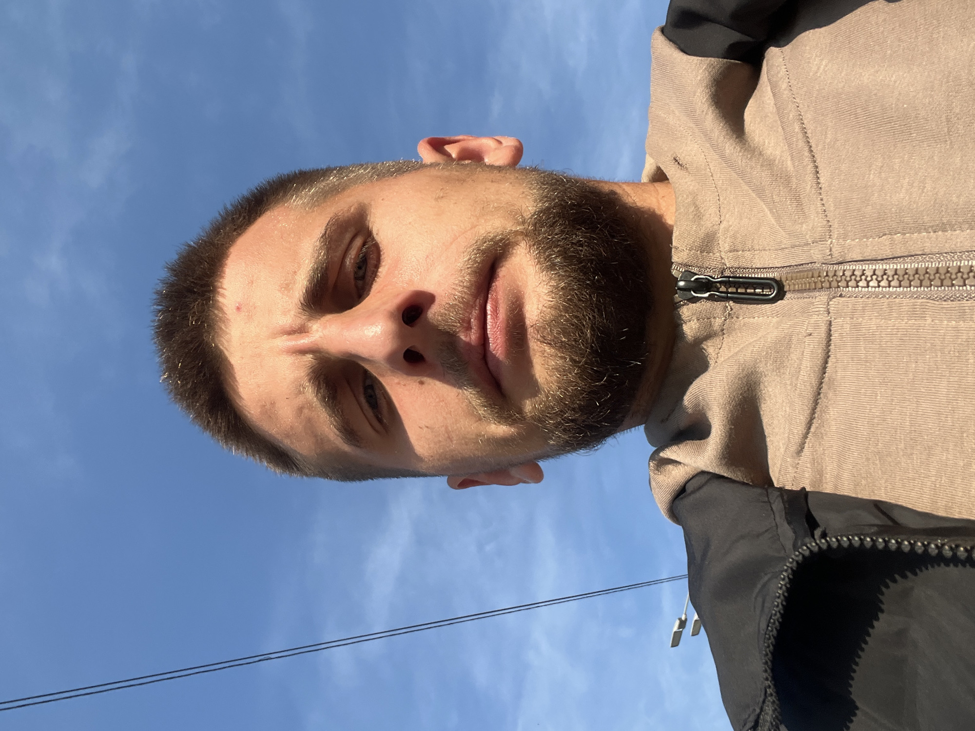 The image is a low-angle, close-up selfie of a young man with short brown hair and a beard, looking directly at the camera. He is wearing a light brown hooded sweatshirt with a black zippered jacket over it. The background is a clear blue sky with some thin, wispy clouds. A dark, thin wire runs diagonally from the upper left corner towards the center of the frame. Two streetlights are visible in the distance, also in the upper left quadrant. The overall impression is that the photo was taken outdoors during the daytime. Without additional context or visual cues within the image, it is impossible to determine the specific location within Moscow, Russia.
