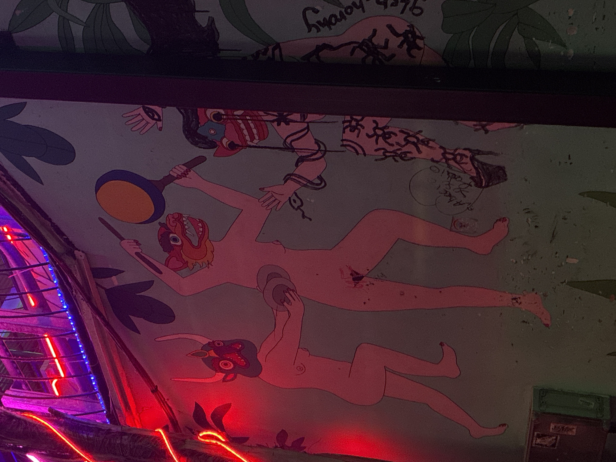 The image shows a ceiling mural in a bar in Tel-Aviv, Israel. The mural depicts nude figures with animal masks, rendered in a cartoonish style, arranged in a scene suggesting a ritual or fantastical gathering. One figure holds a frying pan, while another holds circular objects over a second figure. The top portion of the mural has a character bound in rope that ends in a snake. The room has a neon LED sign in the lower-left corner, giving off red and blue light. A green electrical box is present in the lower-right. The overall environment appears to be dimly lit.
