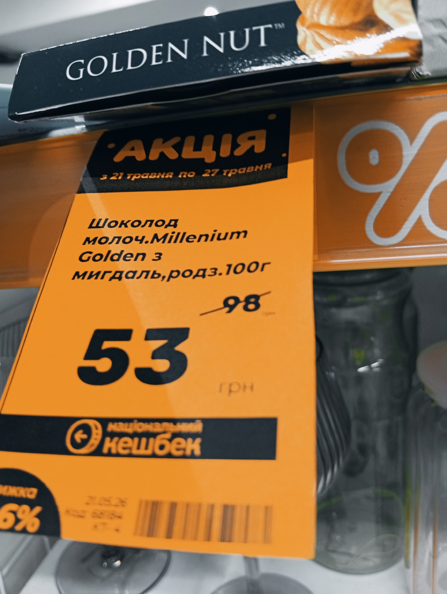 Here is a detailed description of the image:
**Scene:** The image is a close-up shot of a price tag for a chocolate product in what appears to be a retail setting, possibly a supermarket or grocery store.
**Objects:**
* **Price Tag:** The price tag is predominantly orange with black text and graphics. It prominently displays the price of "53" (likely in Ukrainian Hryvnia - Грн). The original price of 98 has been crossed out.
* **Chocolate Product:** The price tag is advertising "Шоколод молоч. Millenium Golden 3 мигдаль, родз.100г" - translated as "Milk chocolate Millenium Golden 3 almond, raisin. 100g".
* **Branding:** The price tag indicates that the product is part of a promotion "АКЦІЯ" - translated as "Promotion" and that the promotion is valid from "3 21 травня по 27 травня" - translated as "from May 21 to May 27".
* **Cashback:** The price tag also includes the logo for a "національний кешбек" - translated as "National Cashback" program.
* **Product Packaging:** Above the price tag, a portion of product packaging is visible with the branding "GOLDEN NUT™".
* **Background Objects:** In the background, through the price tag, there appear to be jars or bottles on a shelf.
**Environment:** The lighting is bright and likely artificial, typical of a retail environment.
**Location:** It is likely to be in Ukraine, given that the currency is Ukrainian Hryvnia and the text on the price tag is in Ukrainian. The brand 'Millennium' is based in Dnipro, Ukraine. It is not possible to determine if the photo was taken in the city of Poltava, Ukraine, without more information.