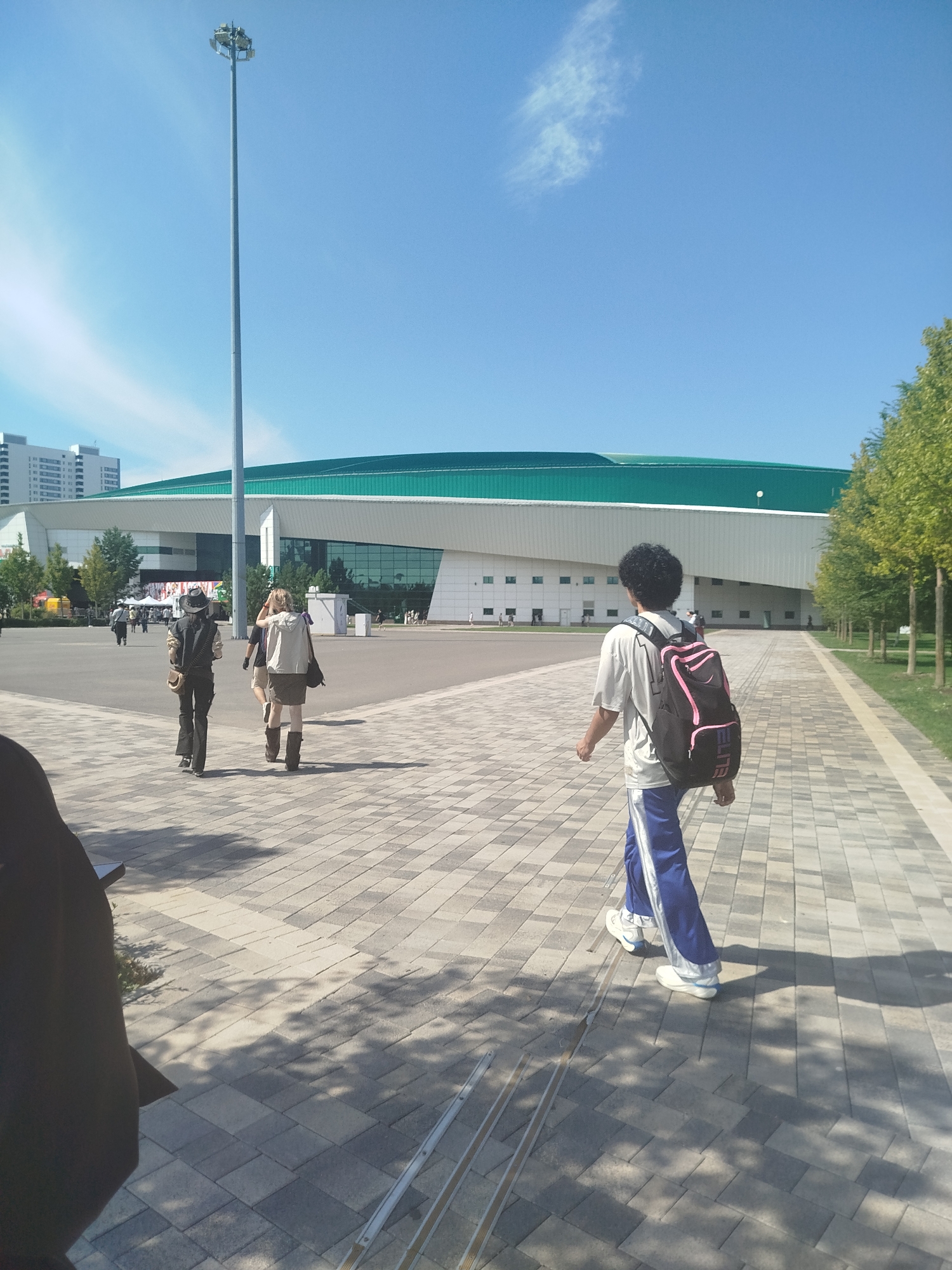 The image shows a wide, paved walkway outside a large, modern building complex with a green, curved roof. The building appears to be a stadium or arena, given its scale and design. A tall lamppost stands prominently to the left of the building. Several people are walking on the walkway. In the foreground, on the right, a person with dark curly hair is walking away from the camera, wearing a light-colored t-shirt and blue track pants with white stripes, carrying a black backpack with pink accents. To the left of this person, another individual is walking away, wearing a light-colored jacket and a dark skirt. Further to the left, a person in a cowboy hat, dark shirt, and pants walks with a crossbody bag and a backpack. The walkway is paved with light-colored rectangular stones. Trees line the right side of the walkway, and shadows from the trees and people are cast on the ground. In the background, a residential building with many windows is visible. The sky is clear and blue with a few wispy clouds. This scene is consistent with the area around the Astana Arena (now known as the Saryarka Velodrome) in Astana, Kazakhstan, a well-known multi-purpose sports venue.