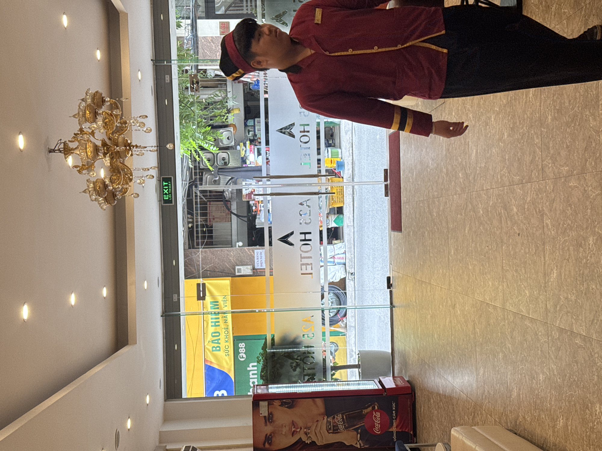 A man in a maroon uniform with yellow trim and a matching cap stands in the foreground of a hotel lobby in Hanoi, Vietnam. He is walking towards the right of the frame. In the background, through a glass door, a street scene is visible, with trees and buildings. A Coca-Cola refrigerator with an image of a woman is to the left of the entrance. A chandelier hangs from the ceiling. The floor is tiled.