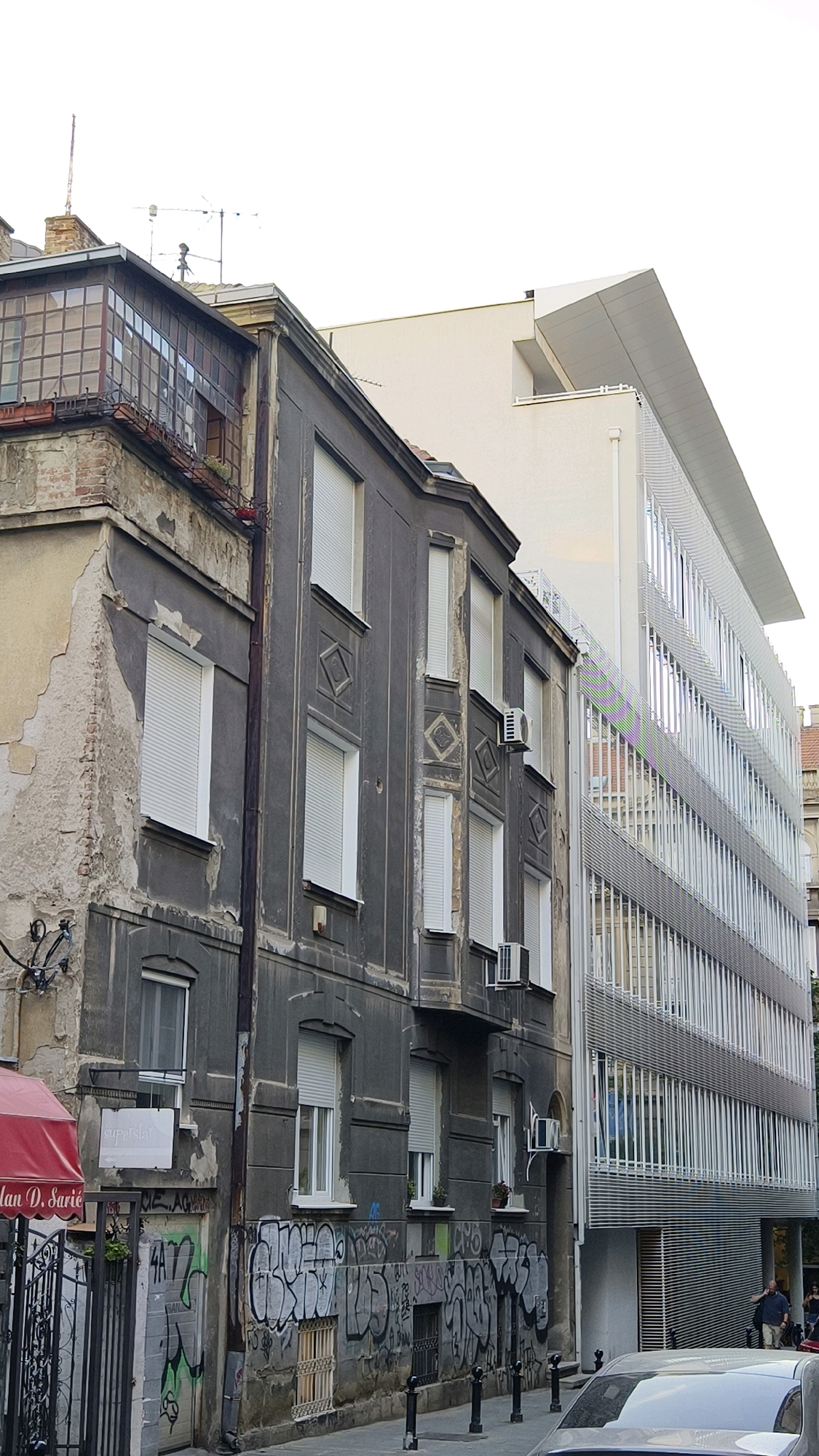 The image depicts a street view in Belgrade, Serbia, showcasing a juxtaposition of old and new architecture. On the left, an older, weathered building with peeling paint and visible brickwork stands tall. It features numerous windows with white roller shutters, some adorned with decorative diamond patterns. The building has a bay window on its upper floors with a grid-like glass facade. Adjacent to this, a modern, white office building with a sleek, geometric design and a facade covered in horizontal metal slats dominates the right side of the frame. The contrast between the crumbling facade of the older structure and the clean lines of the new building is striking. The street level of the older building is covered in graffiti, suggesting a more urban, perhaps gritty, atmosphere. A red awning for "Milan D. Sarić" is visible on the far left. The sky is overcast and white, indicating a cloudy day. In the distance, a few people can be seen walking, and a silver car is parked in the foreground, partially obscuring the view.