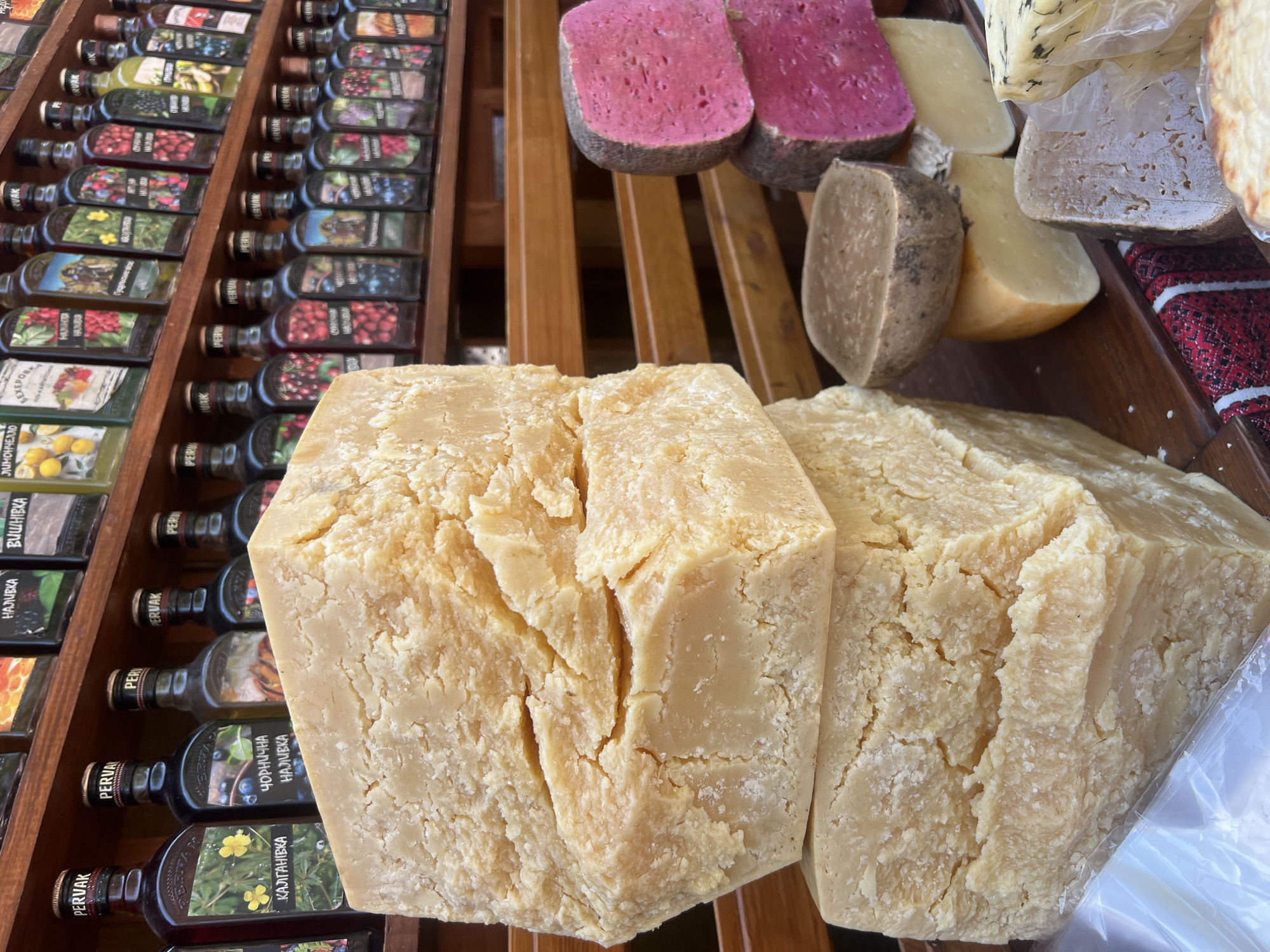 In Yaremche, Ukraine, on a wooden shelf at a market stall, two large blocks of light yellow cheese are stacked in the foreground. To the right of the cheese blocks, several rounds of cheese are displayed, including two with a bright pink interior and grey rind, one smaller round with a brown crust, and a wedge of yellow cheese. Further to the right, more cheese is visible, some wrapped in plastic, with herbs embedded in one variety. In the background, two shelves are filled with numerous bottles of various colored liquids, many labeled with the brand name "PERVAK" and featuring images of fruits and berries. The bottles appear to be tinctures or spirits. The overall scene depicts a food market offering cheese and alcoholic beverages.
