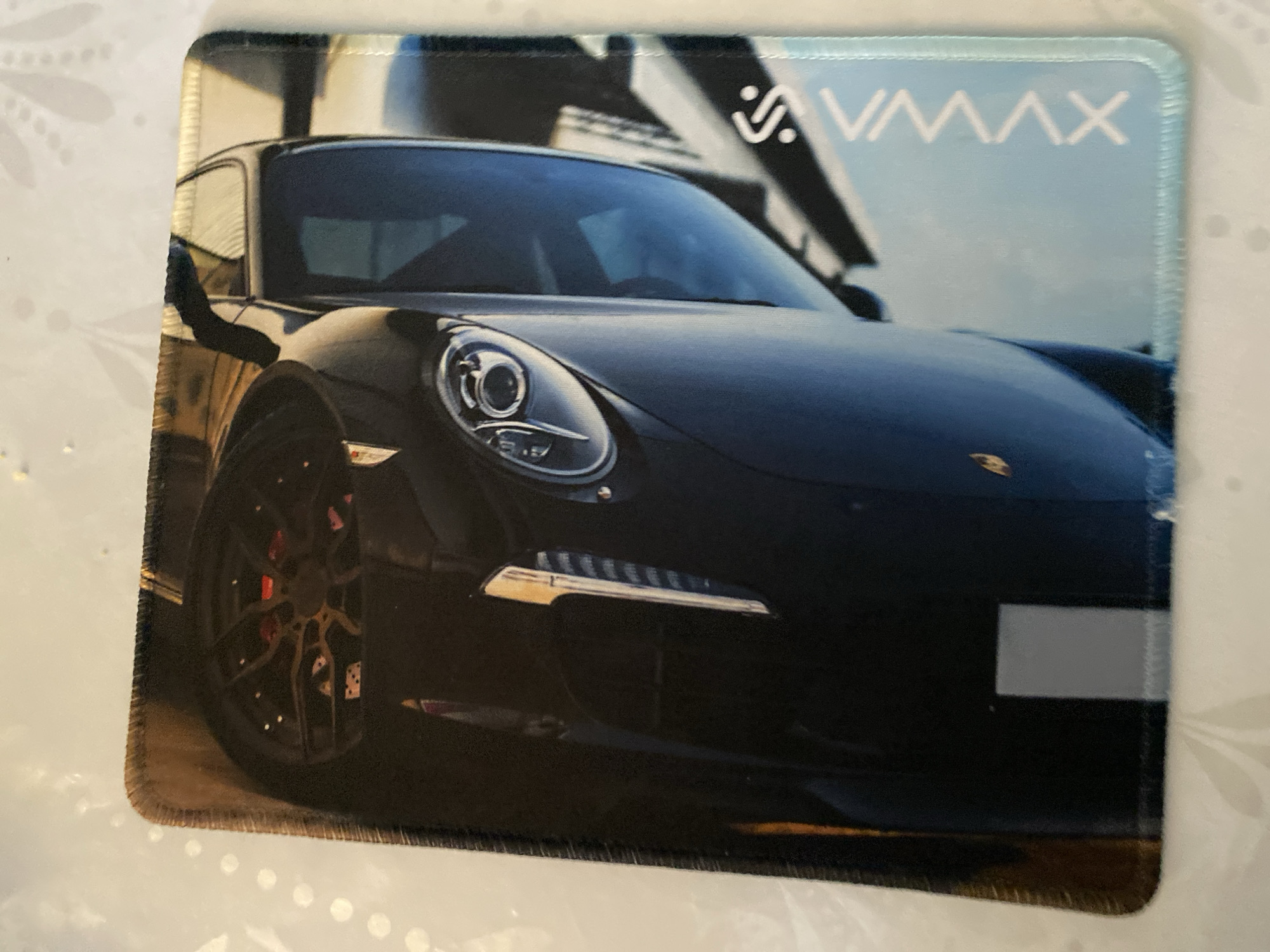 An overhead, slightly angled, full shot captures a close-up view of a dark blue sports car, likely a Porsche, with its front end angled towards the lower left of the frame. The car's headlight, a circular lens with internal detailing, is visible on the right side of the hood. A portion of the car's wheel, featuring red brake calipers and a complex silver spoke design, is visible in the upper left. The car's body appears sleek and polished, reflecting the ambient light. To the right of the car, a portion of another car, or a reflection of the same car, is visible in a lighter blue hue. In the lower right corner of the image, white text reads "VMAX" vertically, with a stylized "S" above it. The image is printed on a rectangular object with a stitched, dark border, resembling a mousepad. The background is a white, textured surface with faint floral-like patterns.