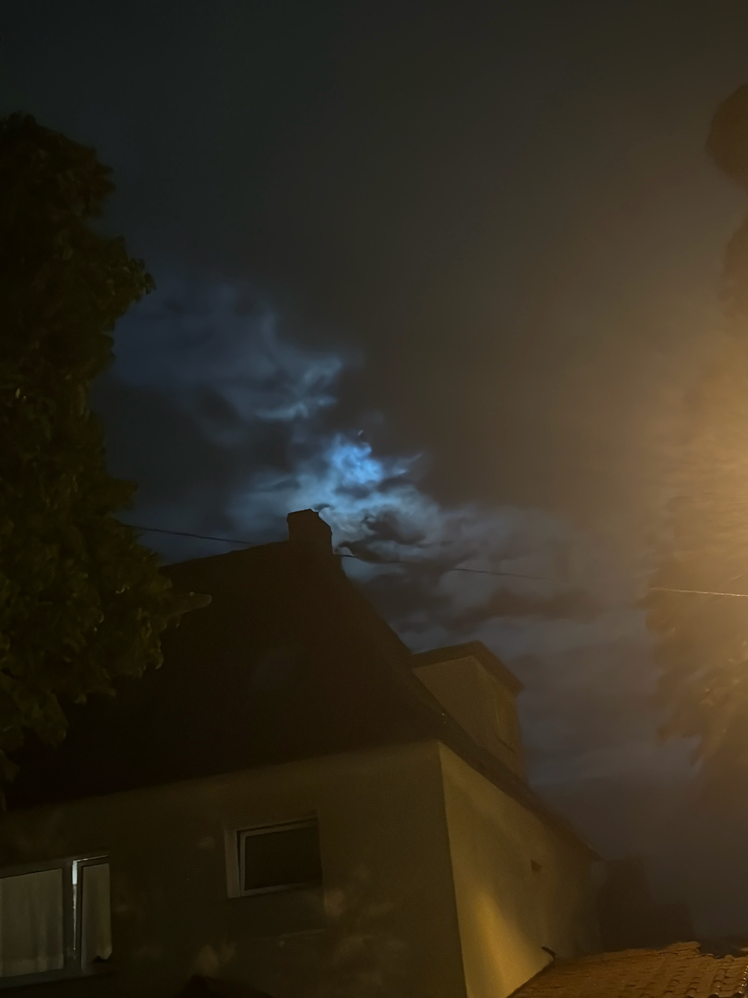 The image depicts a night scene in Szczecin, Poland. A tree is visible on the left, with its leaves dark against the night sky. In the center, the roof of a building, likely a house, is silhouetted against the sky. A chimney stands on the roof. The sky is filled with dramatic, swirling clouds illuminated by a bright, bluish light, suggesting the presence of the moon, possibly partially obscured by the clouds. To the right of the image, a streetlamp casts a warm yellow light on the scene. The lower portion of the image shows the side of a light-colored building with two windows, one of which appears to be open. The overall atmosphere is moody and atmospheric, typical of a cloudy night with moonlight.