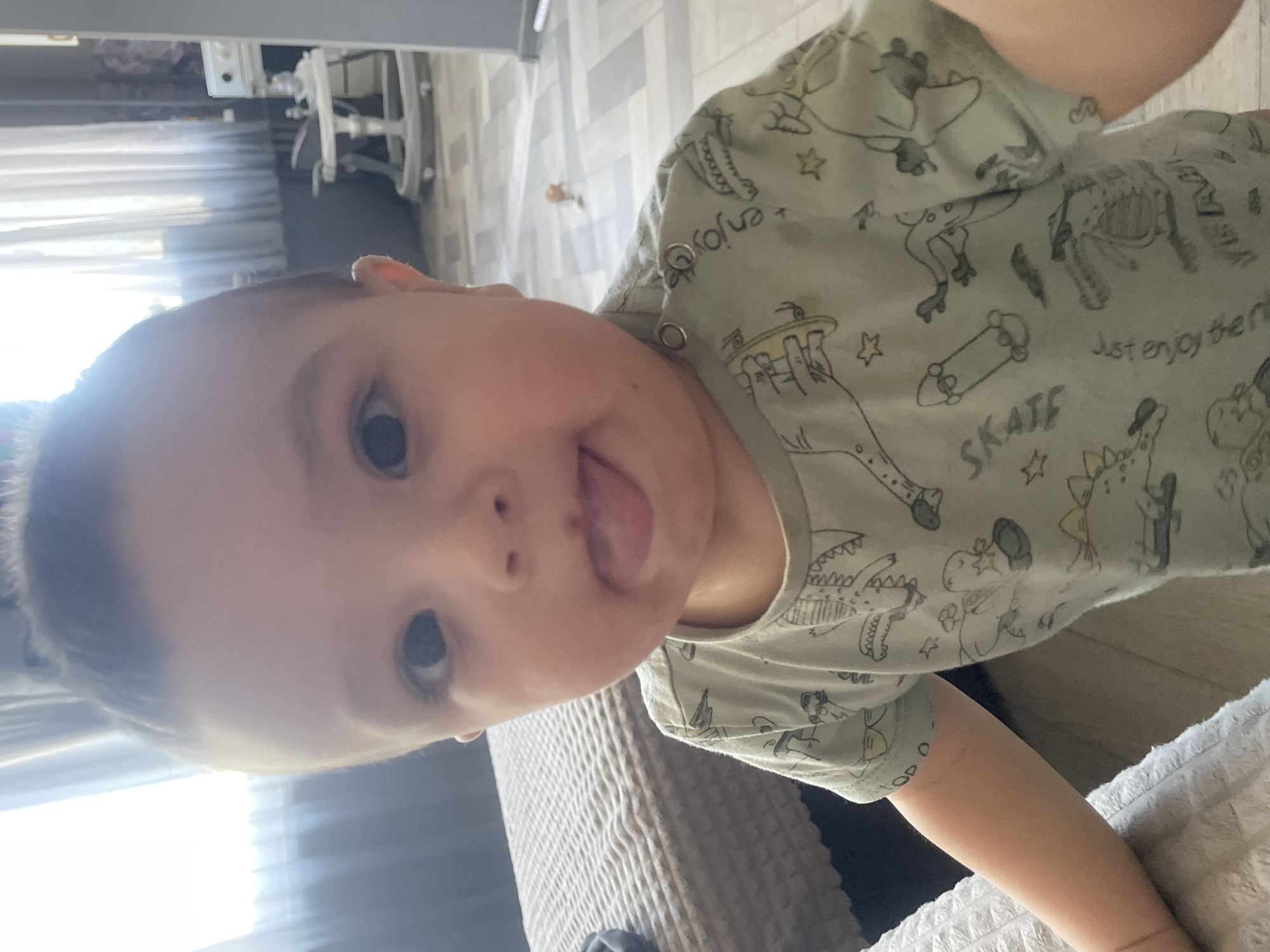 A baby is sticking its tongue out at the camera. The baby is wearing a green shirt with illustrations of skateboards and animals. The background includes a gray couch and striped curtains. The location is Chaikovsky, Russia.