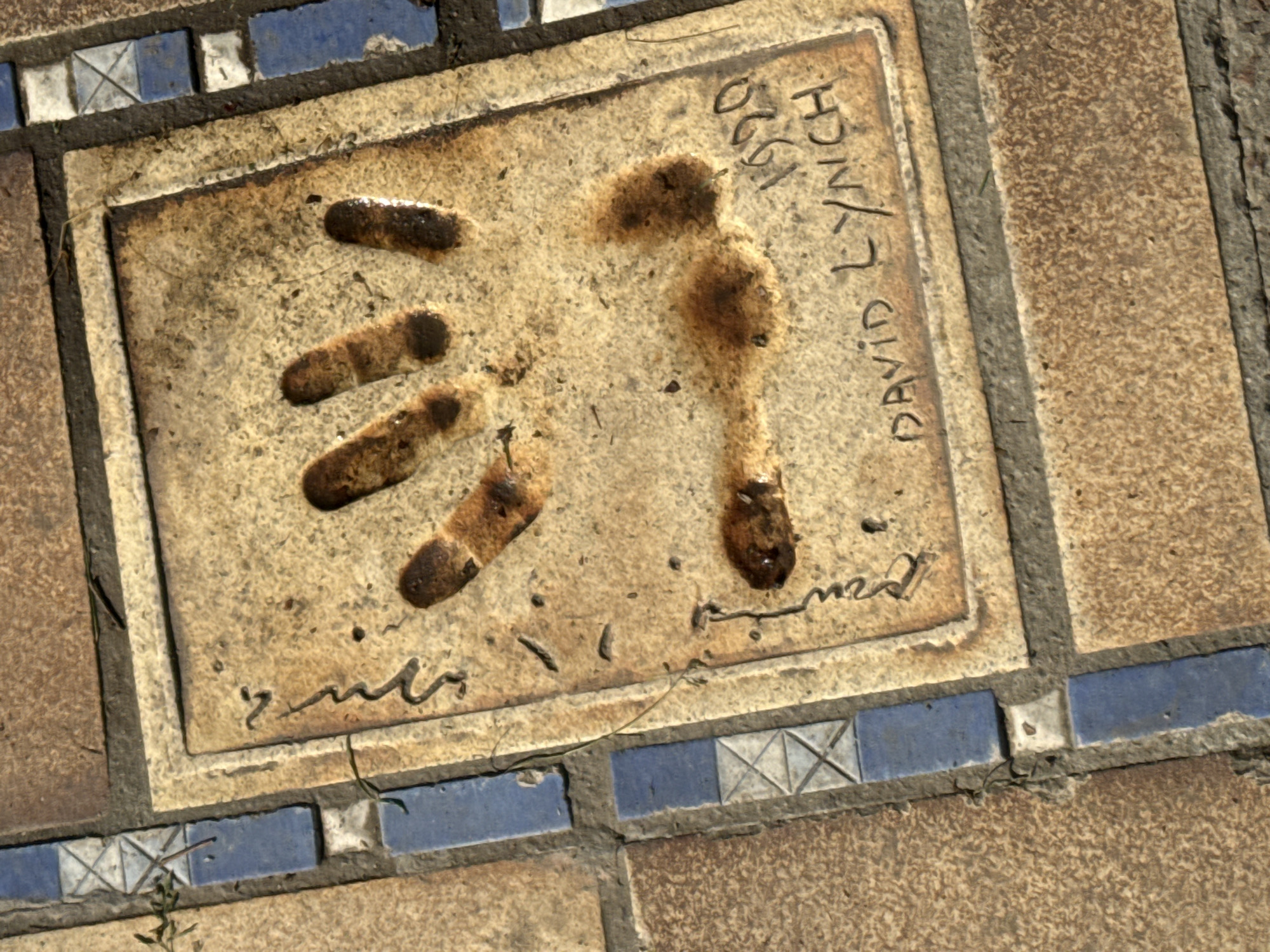 The image shows a rectangular plaque embedded in a tiled surface, featuring the handprint of David Lynch, along with his signature and the year 1990. The plaque, which appears to be aged and slightly soiled, is bordered by blue and white mosaic tiles. The surrounding tiles are larger and have a brown-tan color. This handprint plaque is located in Cannes, France, likely as part of a collection of celebrity handprints, in an area associated with film or entertainment.