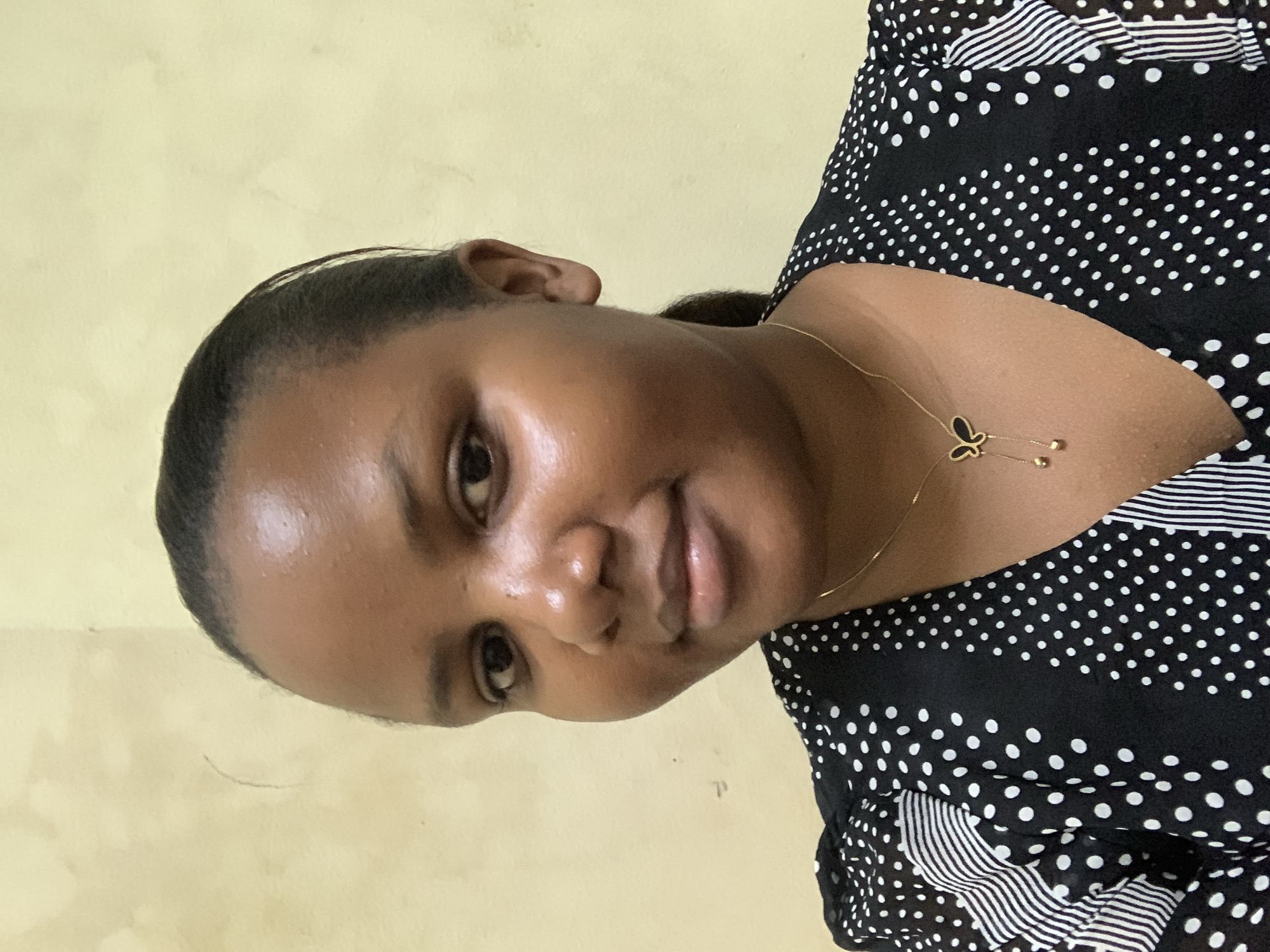 A medium indoor shot presents a woman with dark hair pulled back from her forehead, wearing a black and white polka dot top with striped accents. She wears a gold necklace with a butterfly pendant. The background is a plain yellow wall. It is not possible to determine the location in Dar es-Salaam or any other specifics without additional information.