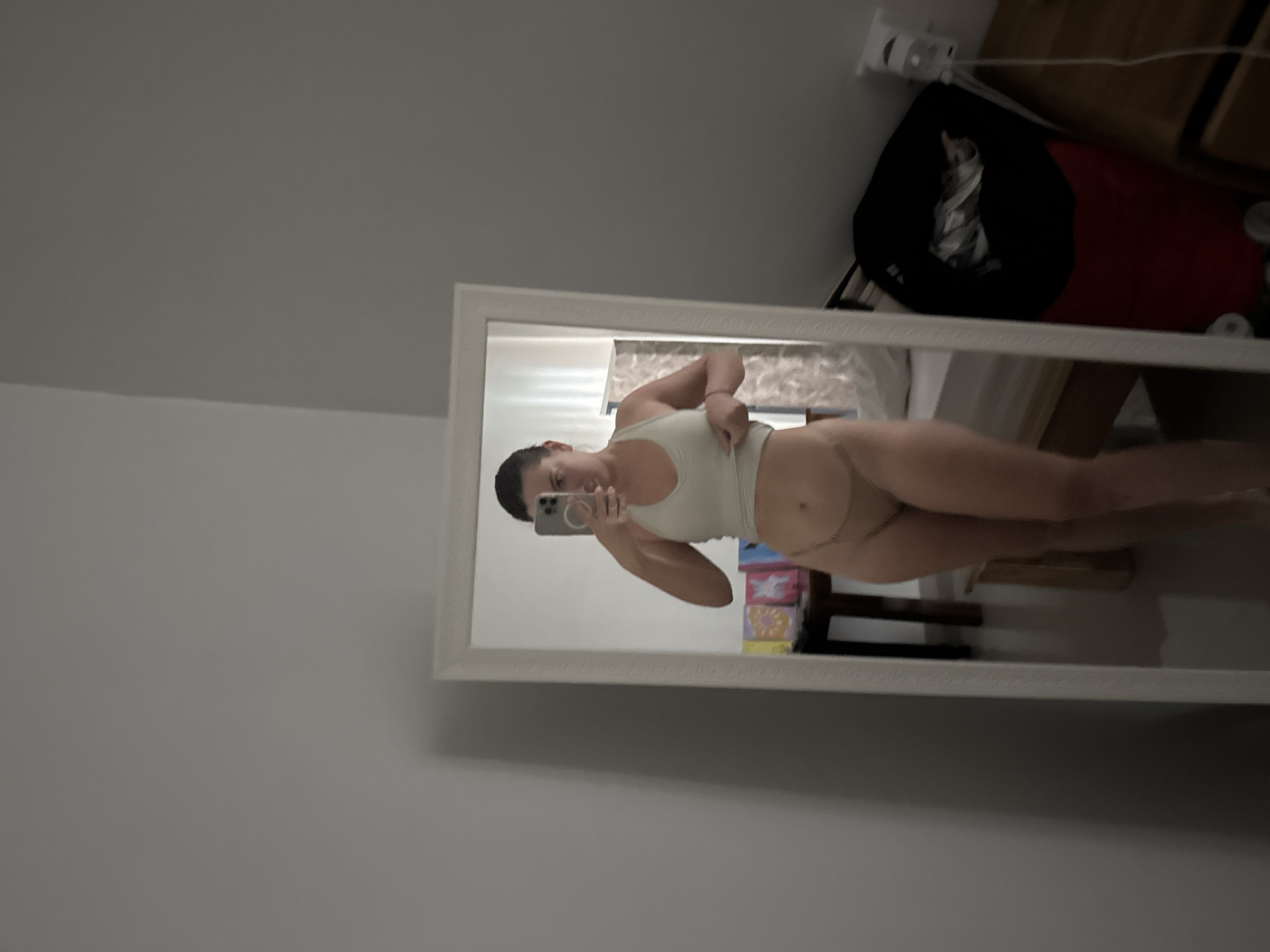 An interior scene depicts a female individual taking a mirror selfie within a bedroom setting, located in Dalung, Indonesia. The individual, appearing in full length, is reflected in a white-framed mirror positioned against a light grey wall. She is observed holding a light-colored smartphone with her left hand, aimed towards the mirror to capture the image. Her right hand is positioned at the lower edge of her light-colored sleeveless crop top, slightly pulling it upwards to expose a portion of her abdomen. She is also wearing light-colored briefs.
The reflection in the mirror reveals elements of the room: to the subject's left, a dark-toned wooden side table holds several colorful, possibly framed, items. To her right, a bed with a white mattress and a light headboard is partially visible. Further to the right of the bed, a black and red bag, appearing to be luggage or a hamper, is situated next to a wooden cabinet or dresser. A white power strip with associated cabling is mounted on the wall above this furniture. The room features light-colored walls and a light-toned tiled floor. The ambient lighting is uniform.
