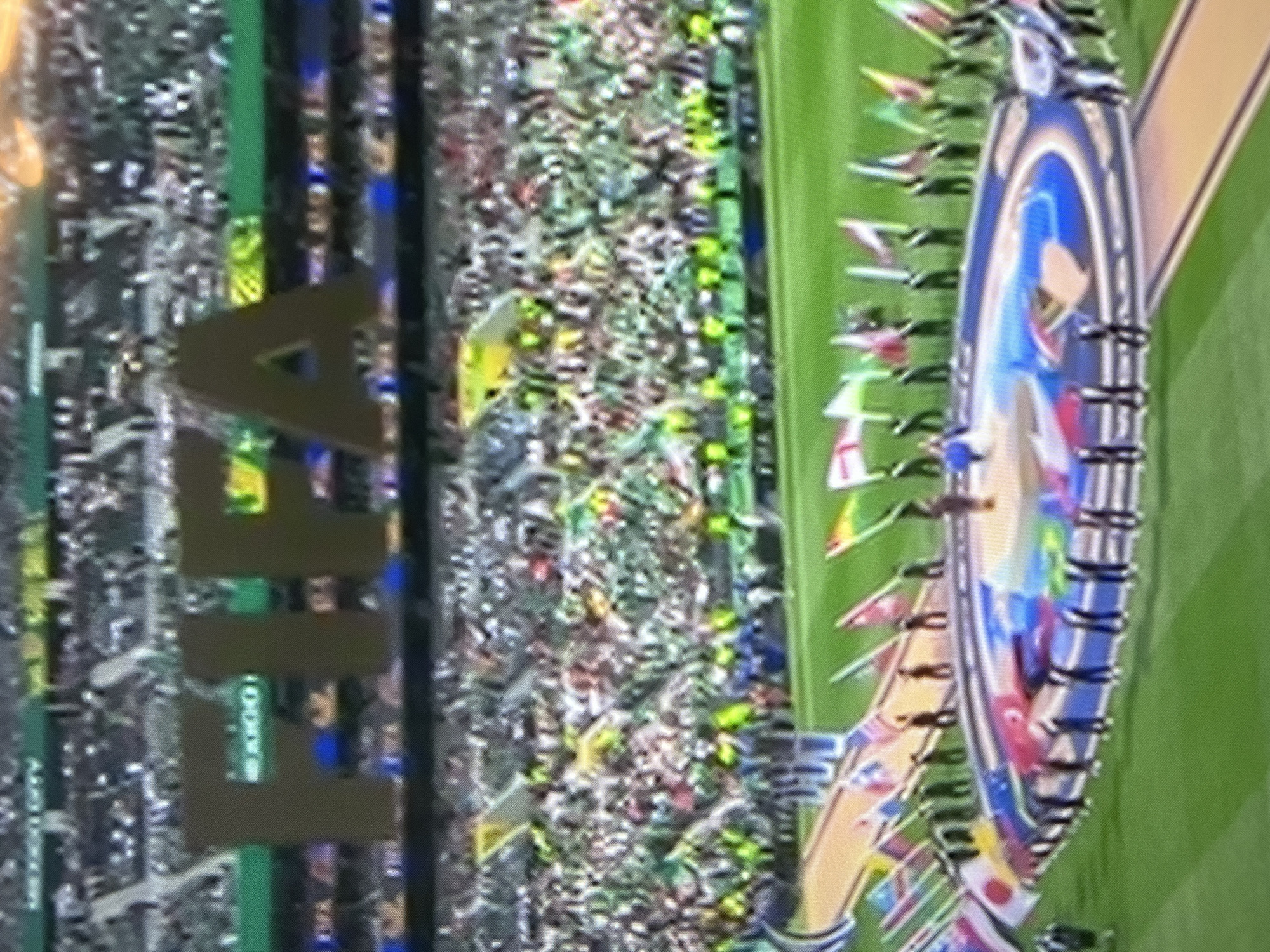 A wide-angle, slightly blurred, eye-level shot captures a stadium event, likely an opening ceremony. In the background, large, brown, block letters spell "FIFA" across a green backdrop. The stadium is filled with a large crowd, many of whom are wearing green and white, waving flags. In the foreground, a circular stage area is decorated with the flags of various nations arranged in a semi-circle. Several individuals, appearing to be performers or organizers, are standing on and around the stage, some holding flags. The overall atmosphere suggests a significant sporting or cultural event.