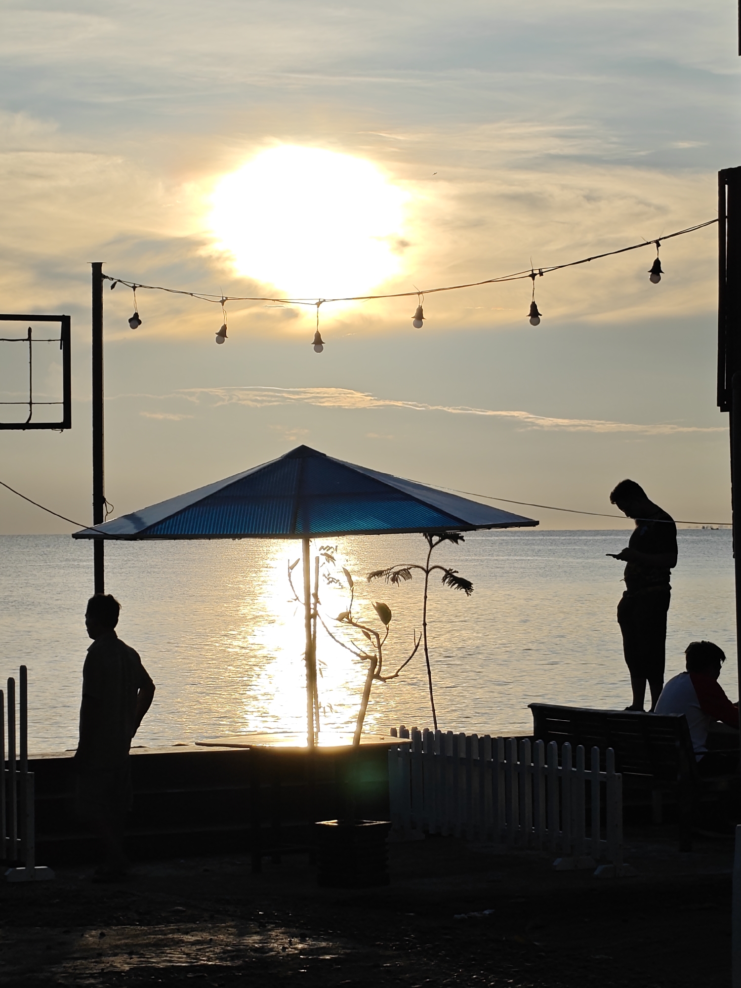 The image captures a scene on a beach in Lhokseumawe, Indonesia, during sunset. The sun is setting over the water, casting a bright glare on the sea and sky. A string of lights hangs overhead, and a blue parasol stands in the middle ground. Several people are visible in silhouette. One person stands alone, looking at their phone. Another person sits on a bench. A white picket fence is visible in the foreground.