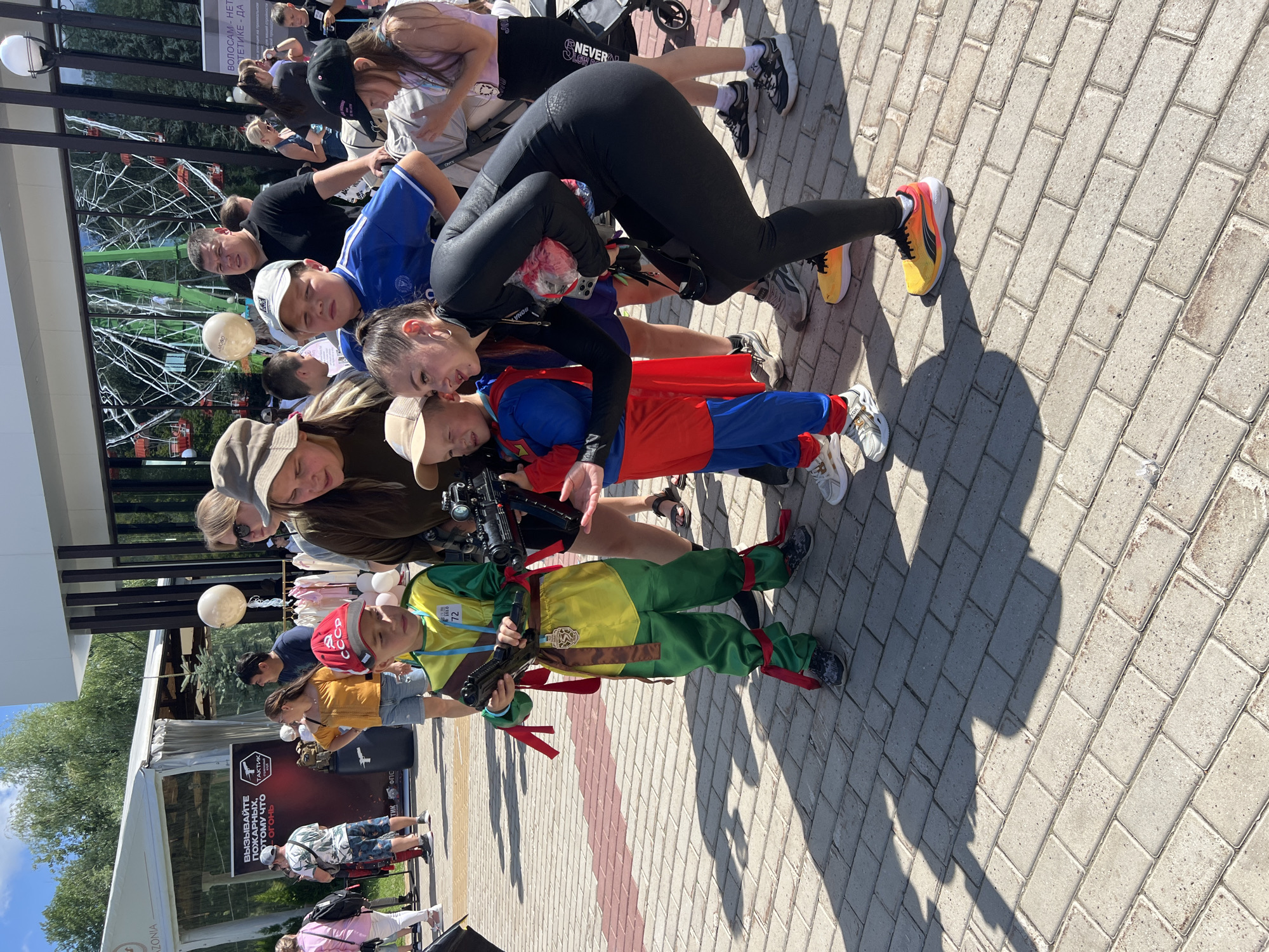 A group of people, including several children in costumes, are gathered on a paved outdoor area, likely a promenade or public square, in Cheboksary, Russia. In the background, a large building with a modern facade is visible, along with trees and a ferris wheel. One child is dressed as a ninja, holding a toy gun. Another child is dressed as Superman. An adult woman in a black catsuit is interacting with the children, also holding a toy gun. Other adults and children are observing the scene. The ground is paved with interlocking brick-like stones. The overall atmosphere appears to be a public event or festival.