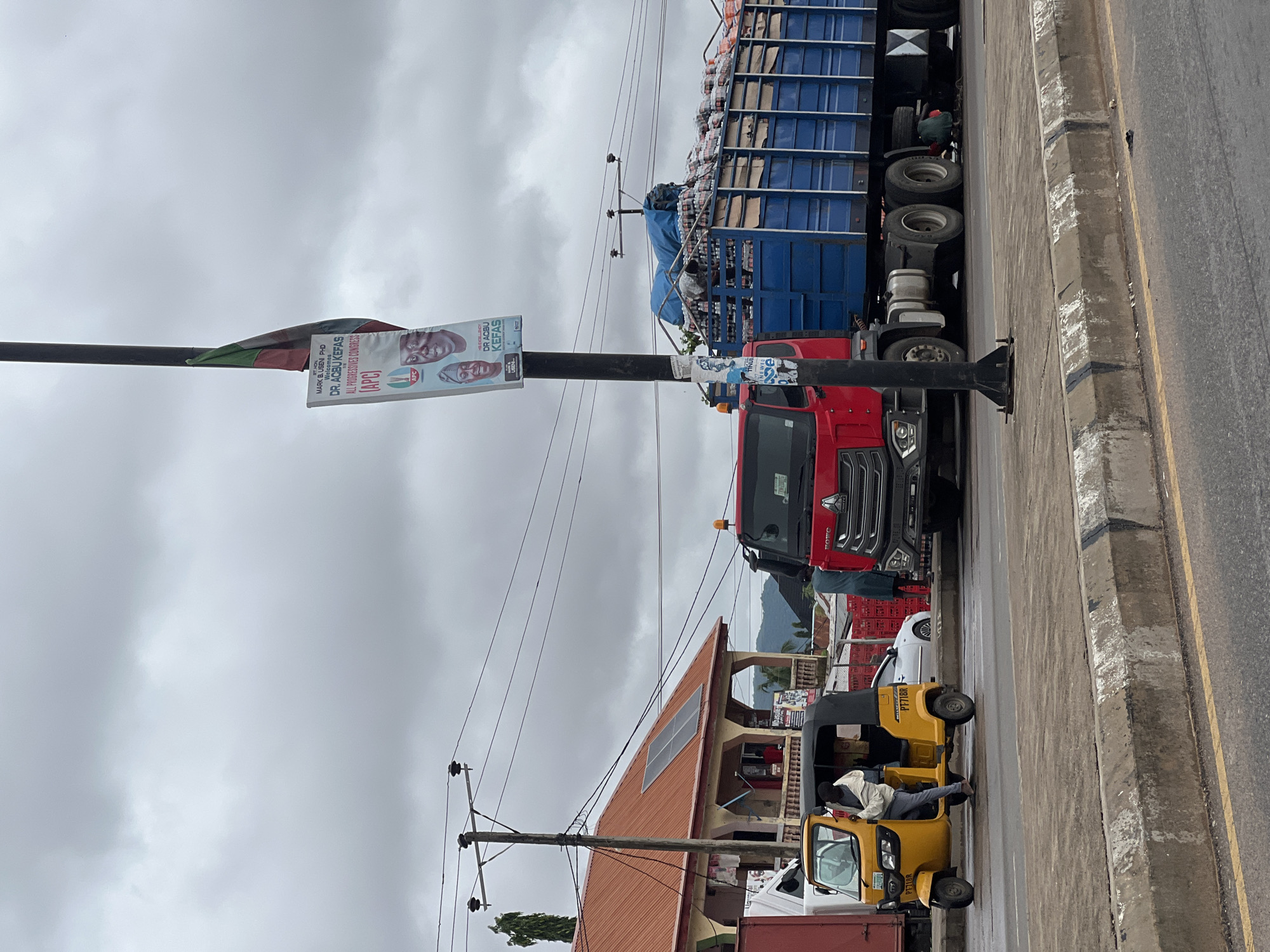 In Jalingo, Nigeria, a red truck with a blue trailer loaded with bottled drinks is parked on a road under an overcast sky. The truck is a HOWO model. A yellow tricycle, also known as an "okada" or "keke," is stopped next to the truck, with a man in a light-colored jacket standing next to it. A white sedan is parked behind the tricycle.
A tall pole stands next to the truck, displaying a political banner with images of two men and the text "DR. AGBU KEFAS." A Nigerian flag is partially visible on the pole.
To the left of the road, a building with an orange roof and solar panels is visible. The building has a covered walkway with columns. Power lines are strung across the scene, supported by wooden poles. The road has a concrete median with yellow and white markings. The overall scene suggests a commercial or transportation hub in the city.