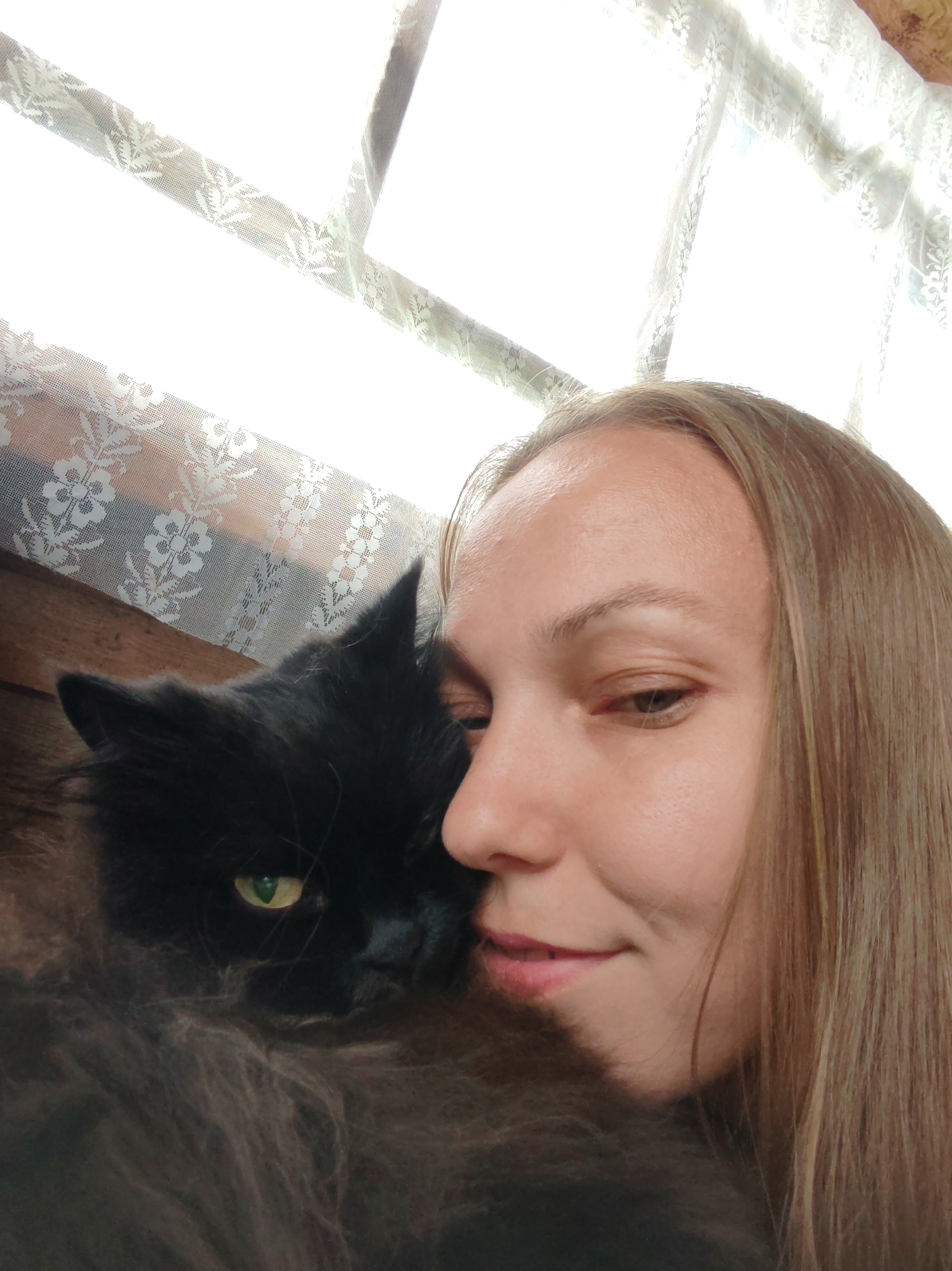 A close-up, slightly overhead shot shows a woman and a black cat nestled together. The woman's face is on the right side of the frame, with her eyes closed or looking down. She has light brown hair that falls around her face and shoulders. She is smiling slightly. The cat is on the left, with its face pressed against the woman's. The cat is black with bright green eyes. It has very fluffy fur. They appear to be indoors, with a white lace curtain visible in the background. The image is taken from a low angle looking up towards the curtain.