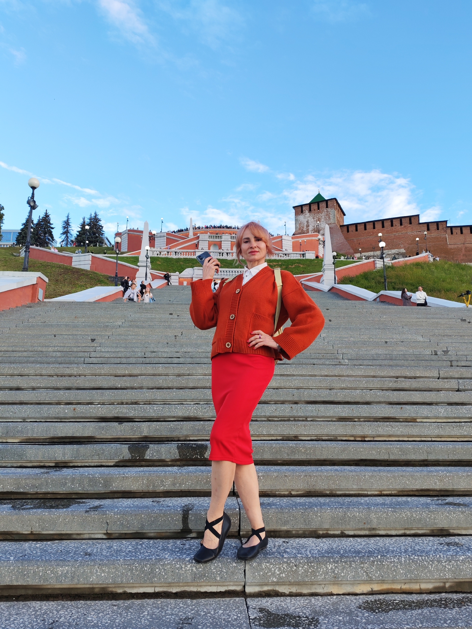 A woman with short, pink-hued hair stands on a wide, grey stone staircase, facing the camera. She is wearing an orange knitted cardigan over a white collared shirt and a form-fitting red midi skirt. Her black ballet flats have an criss-cross strap design. She holds a smartphone in her right hand, which is angled towards her face as if taking a selfie. Her left hand rests on her hip. She is smiling slightly.
Behind her, the staircase continues upwards, bordered by bright orange walls and grassy slopes. Further in the background, a large red brick fortress wall with a green-capped tower stands against a bright blue sky with scattered white clouds. The scene appears to be in the late afternoon or early evening, judging by the lighting. The setting is likely a public landmark or historical site, given the architecture and the presence of other people scattered on the stairs and in the background. There are lampposts visible along the staircase and on the fortress wall. No visible text is present in the image.