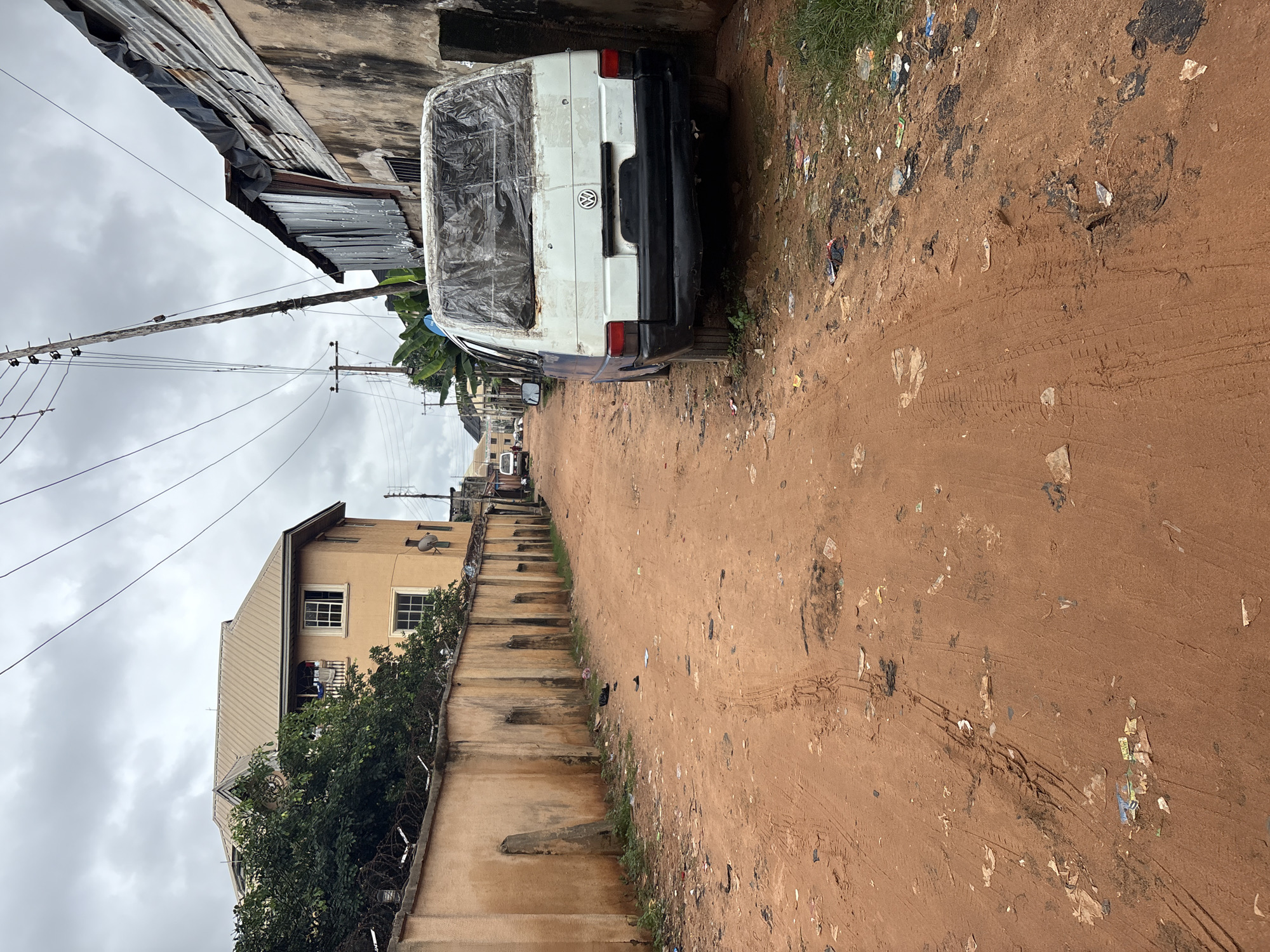 The image depicts a dirt road in Nkpor, Nigeria, characterized by red earth and scattered debris, including plastic waste and tire tracks. On the right side of the road, a white Volkswagen van is parked, covered with a clear plastic sheet. The van's rear window is also covered with plastic. Adjacent to the van is a wall constructed from corrugated metal sheeting attached to a weathered, possibly mud or concrete, structure. On the left side of the road, a tall, light-colored concrete wall runs along the property line, topped with barbed wire. Behind this wall, a multi-story building with a metal roof and tan-colored walls is visible, surrounded by lush green trees. Overhead, electrical wires crisscross the cloudy sky, supported by wooden utility poles. The overall scene suggests a less-developed urban environment with signs of disrepair and informal construction.