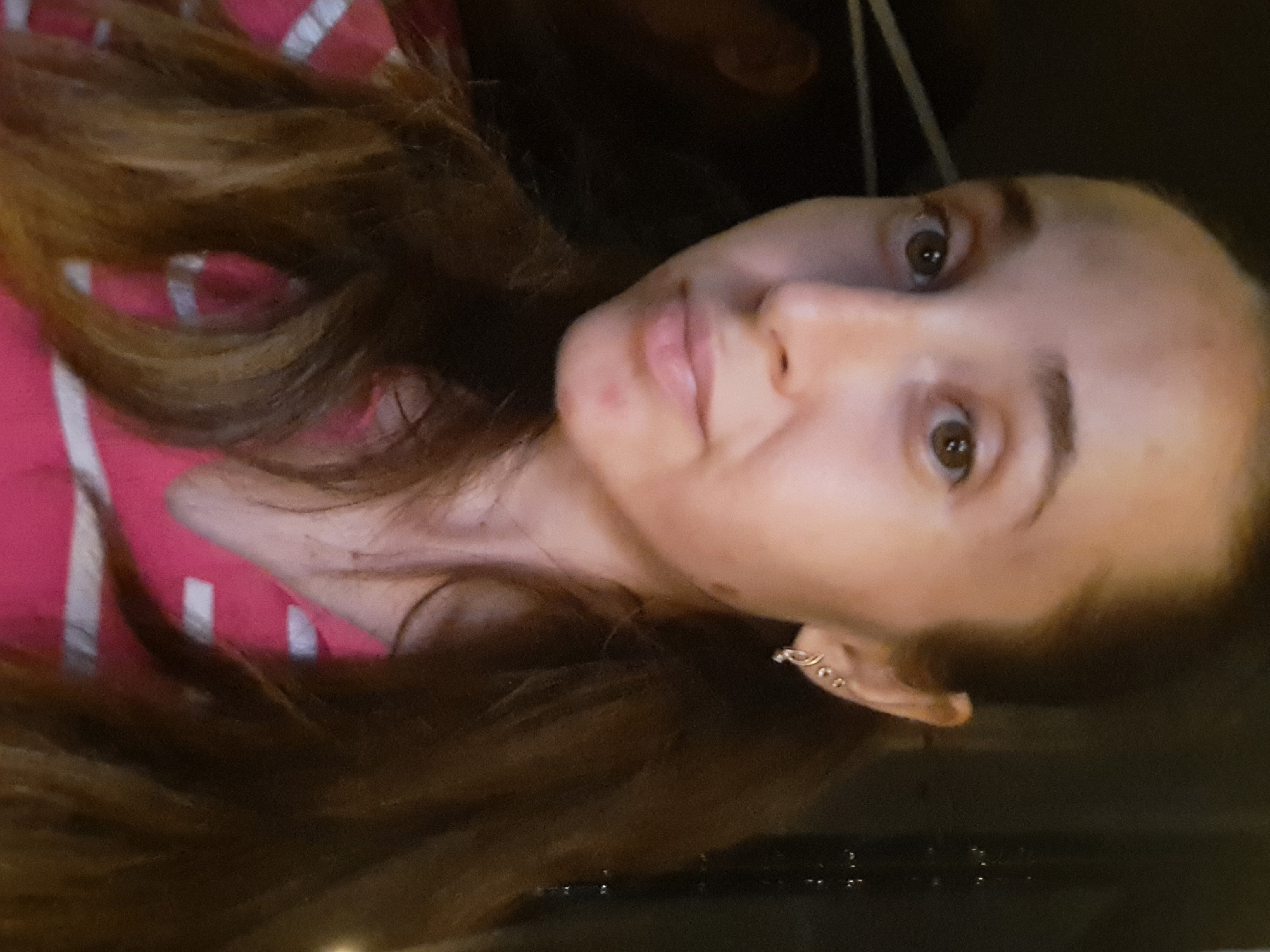 The image shows a medium close-up of a young woman with brown hair, wearing a pink and white striped shirt. She has light brown eyes and appears to have some acne on her chin. She is wearing earrings in her left ear. The background is dark and out of focus, with some faint vertical lines that might suggest a textured surface or reflections. The image does not contain any information that would allow for the identification of a specific location within the city of Vypasne, Ukraine.