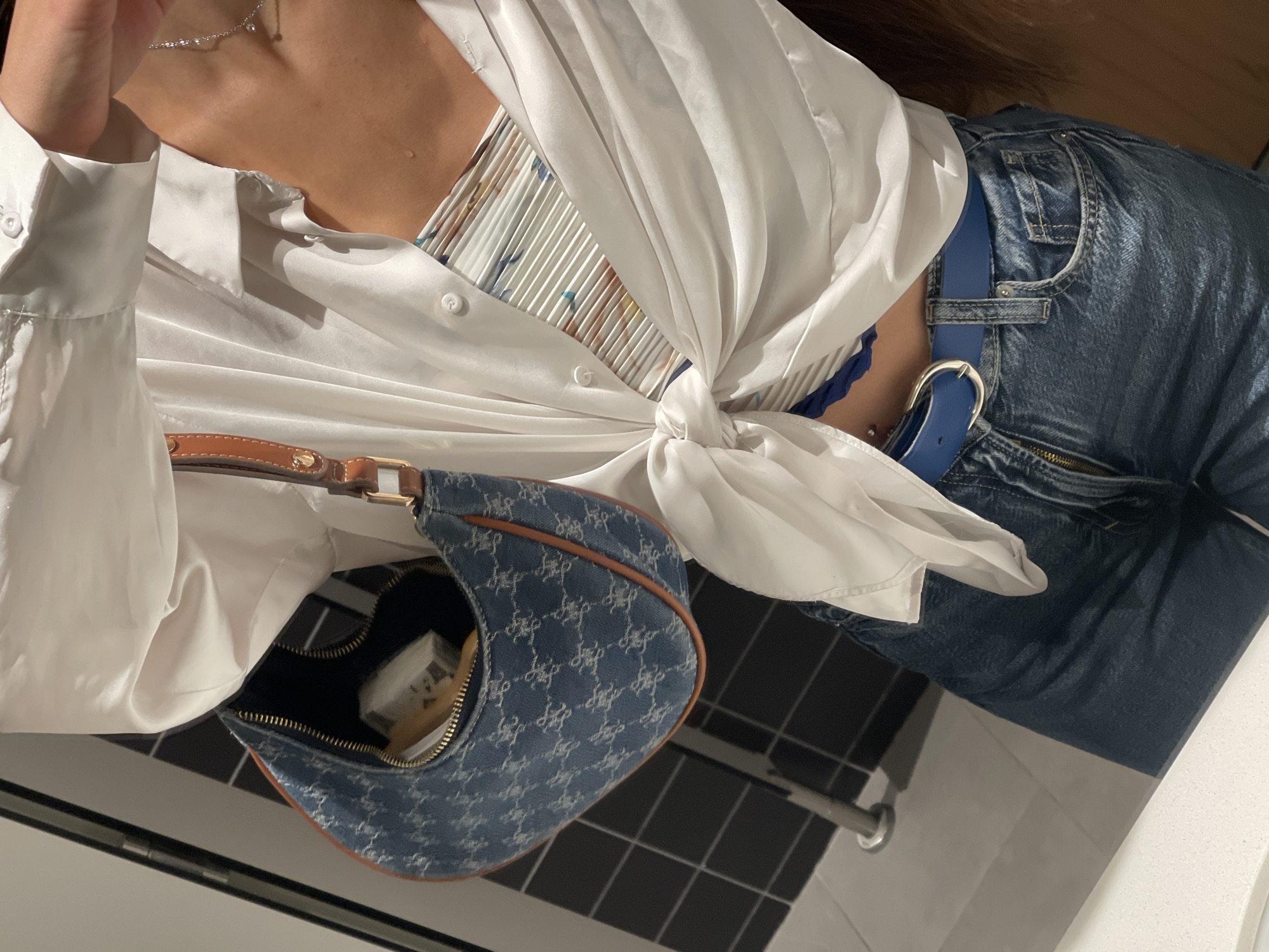 A person is shown from the chest down, wearing a white satin shirt knotted at the waist, revealing a patterned bralette underneath. They are also wearing dark wash denim jeans with a blue belt. A small blue denim purse with brown trim is slung over their left shoulder. A silver necklace is visible around their neck. The person appears to be indoors, standing in front of a tiled wall and floor. The location is not identifiable as being specifically in València, Spain.