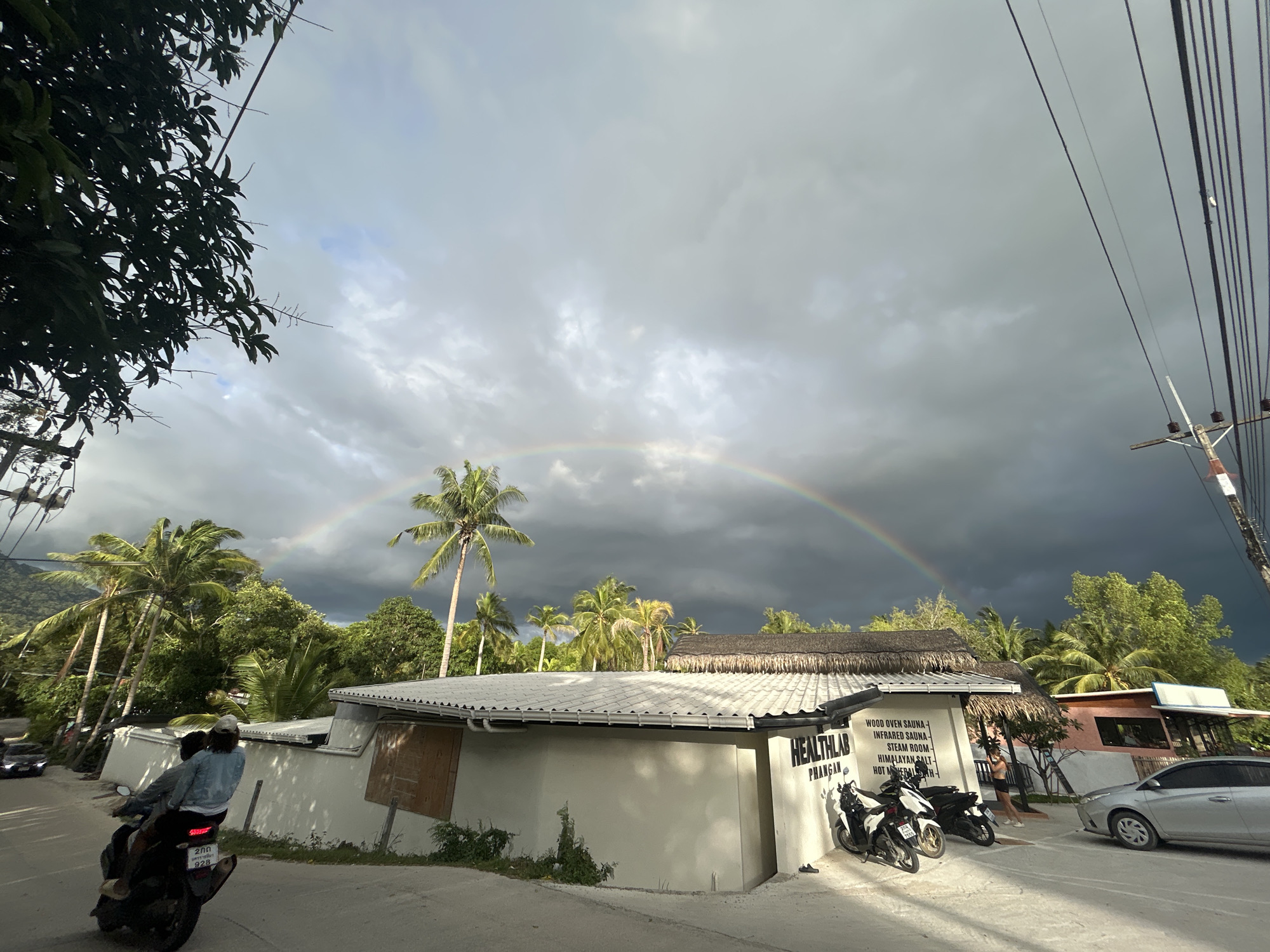 The photograph captures a street scene in Baan Hin Kong, Thailand, under a sky filled with dark, ominous clouds, contrasting with a vibrant rainbow stretching across the horizon. In the foreground, a motorcycle with two people on it is positioned on the left side of the road. The license plate of the motorcycle has the Thai script "200" and the number "928" visible.
To the right, a white building with a thatched roof section houses a business called "HEALTHLAB PHANGAN." A sign lists "WOOD OVEN SAUNA," "INFRARED SAUNA," "STEAM ROOM," "HIMALAYAN SALT," and "HOT MINERAL..." Two motorcycles are parked in front of the building. A silver car is parked nearby.
The backdrop is filled with lush tropical vegetation, including numerous palm trees. Electrical power lines run along the right side of the image, adding a sense of modern infrastructure to the tropical environment.
