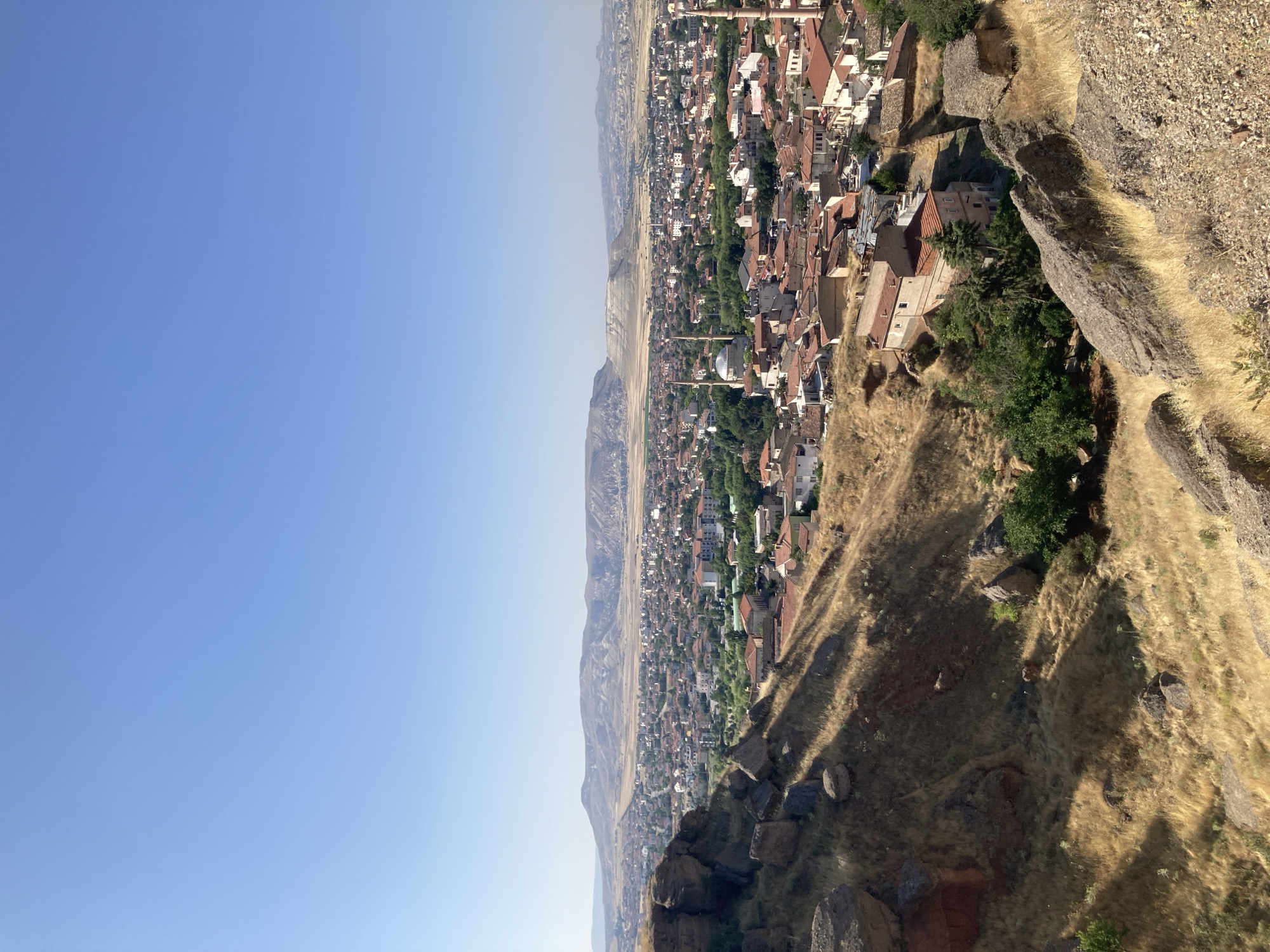 The image shows a panoramic view of the city of Avanos İlçe Merkezi, Turkey, from a hilltop overlooking the city. The foreground is dominated by a rocky, dry hillside covered in golden grass, with large boulders scattered across the terrain. The middle ground reveals the city itself, characterized by a dense arrangement of buildings with red-tiled roofs, interspersed with patches of greenery. Several minarets are visible, indicating the presence of mosques. In the background, a range of low, arid mountains stretches across the horizon under a clear blue sky. The overall impression is one of a sun-drenched, dry landscape with a densely populated urban area nestled within it. There are no people or animals visible in the image.
