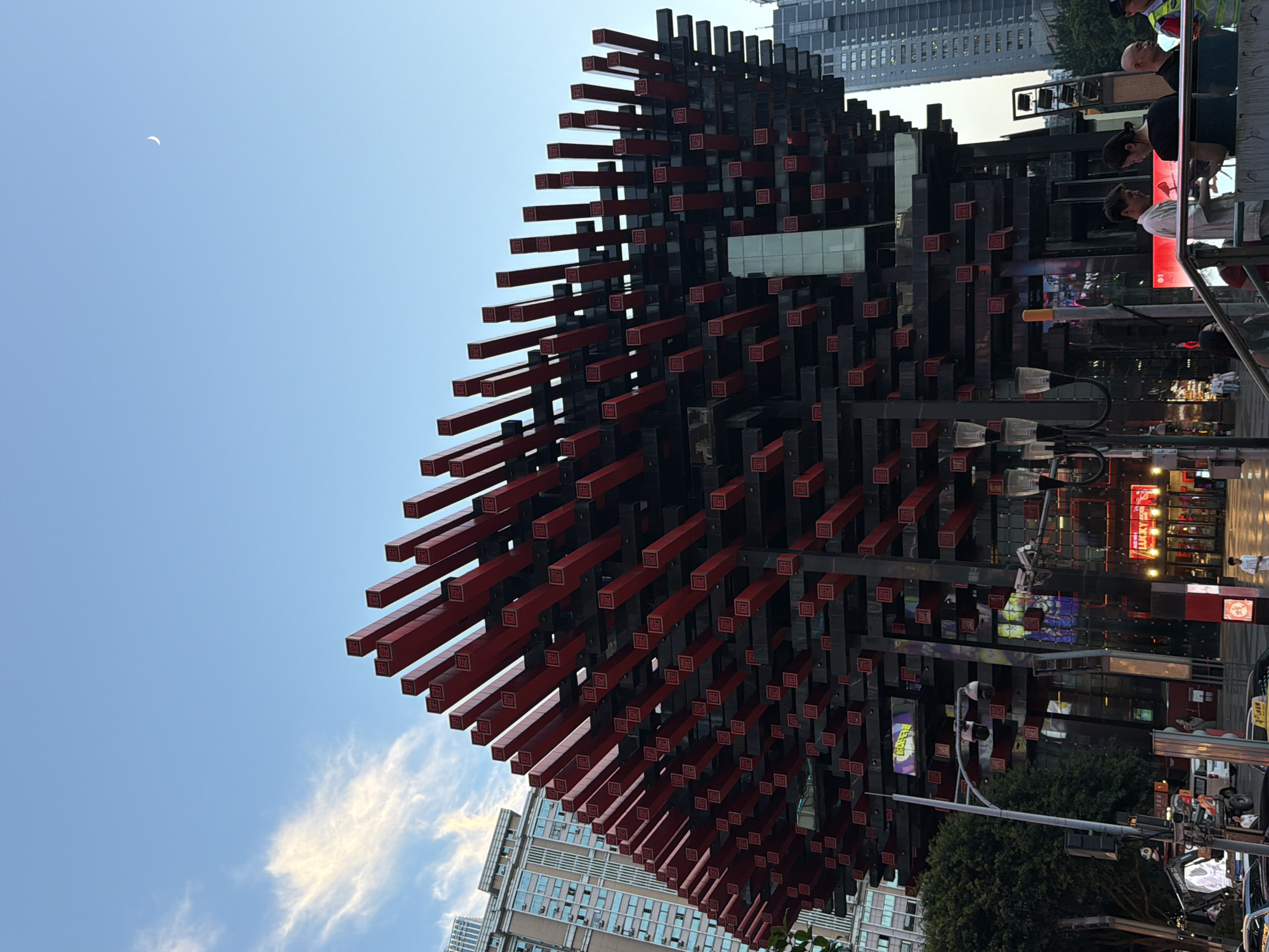 This is a vertical, eye-level shot of a modern building with a distinctive facade featuring numerous protruding red and black rectangular elements. The building appears to be in an urban setting, with other tall buildings visible in the background. The sky is clear blue with a few wispy clouds. A crescent moon is visible in the upper left corner of the frame.
Several people are visible in the foreground and on a higher level of the building, suggesting it might be a public space or commercial establishment. Some are looking at their phones, while others are interacting with each other. The time of day appears to be daytime, with bright sunlight casting shadows. The overall atmosphere seems casual and urban.
The facade of the building is the most prominent feature. The red elements have a repeating Chinese character or symbol on them. Signs with Chinese characters are also visible in the background, indicating the location is in China. The overall impression is one of contemporary urban architecture with a unique aesthetic.