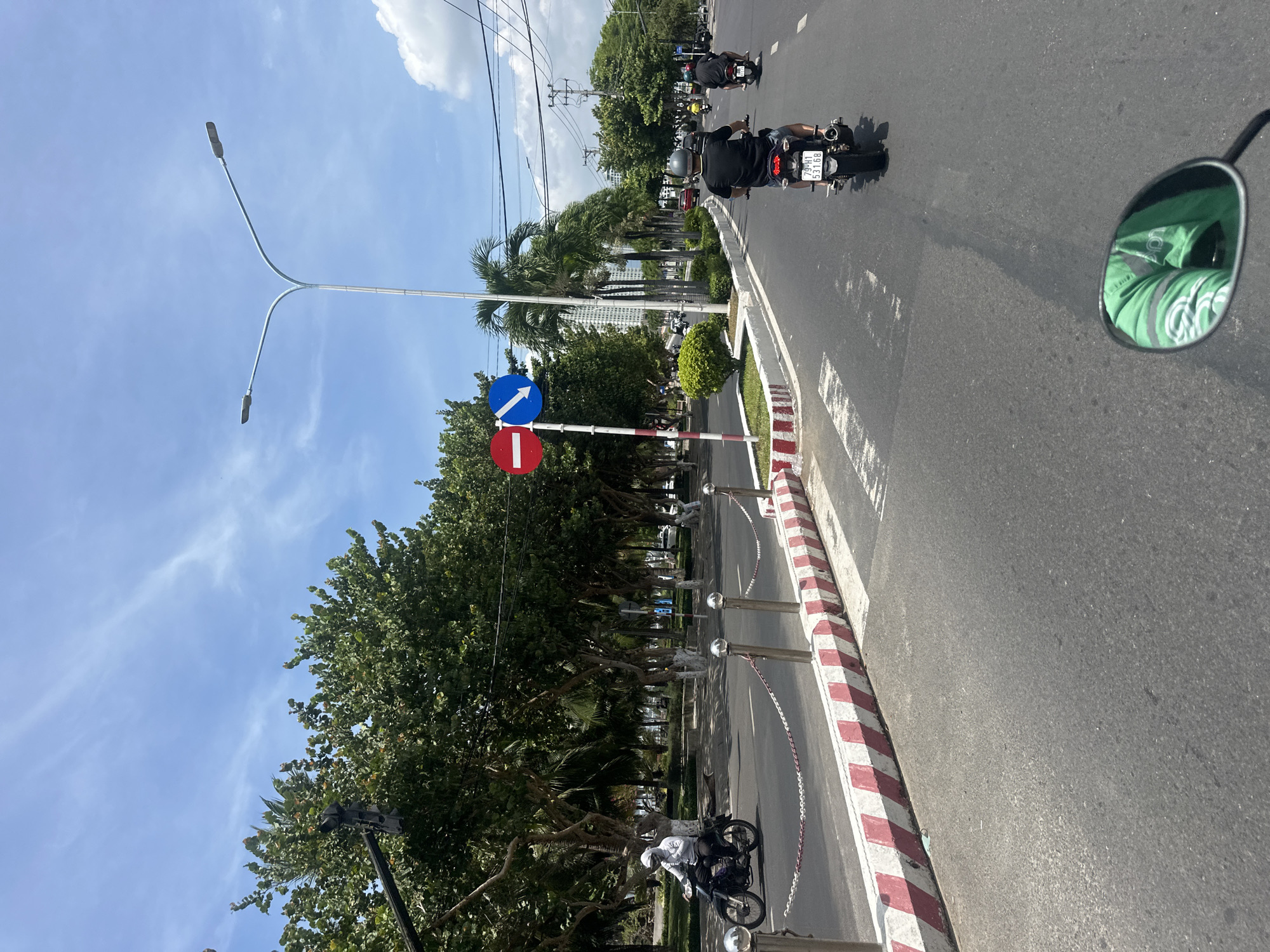 A motorcycle travels along a paved road in Nha Trang, Vietnam. The road is lined with trees and a pedestrian walkway with striped barriers. Traffic signs indicate a "no entry" zone and a mandatory direction. Other motorcycles and cars are visible in the background. The sky is blue with scattered clouds.