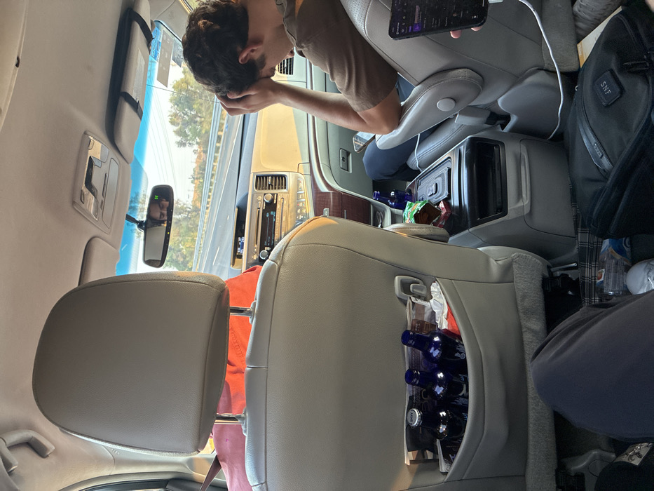 The image depicts the interior of a moving vehicle, identified as being in the vicinity of Santa Ana Xalmimilulco, Mexico. The perspective is from a rear passenger seat looking forward.
In the foreground, the back of a light-colored headrest from the seat directly ahead is visible. The seat-back pocket contains three dark blue bottles, one identified as "Skyy," along with crumpled white material. Below, the legs of a passenger in dark pants and white shoes are partially visible.
To the right, a male passenger in the front seat, wearing a brown shirt and dark pants, is observed with his head down, hands clasped over his head or forehead, suggesting a state of rest, discomfort, or contemplation. Adjacent to him, on the center console, are several snack bags and another dark blue bottle. A white charging cable extends from the console towards the rear right. On the floor to the bottom right, a black bag with "SNF" initials and a plaid object are present. A hand holding a black smartphone displaying a purple-themed music application is partially visible in the lower right.
The vehicle's dashboard and infotainment system are visible between the front seats. The rearview mirror reflects the face of the male driver and displays a green digital "95," indicating speed or a road marker. Outside the windshield, a multi-lane highway with concrete barriers and roadside vegetation is visible under daylight conditions, possibly late afternoon.