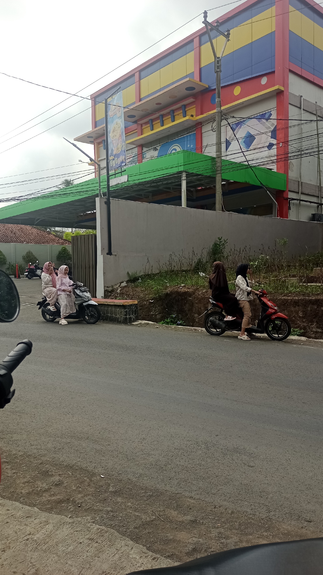 The image captures a street scene in Cikalong, Indonesia, featuring a building with colorful facade and a green awning extending from its front. Two individuals are riding on a motorcycle on the right side of the road, with one person driving and the other as a passenger. On the left side of the road, another motorcycle carries two passengers, both dressed in light-colored clothing and headscarves. The road surface is paved, and there is vegetation and a retaining wall on the right side of the street. A utility pole with wires is visible in the background, near the building. The sky is overcast, suggesting a cloudy day.