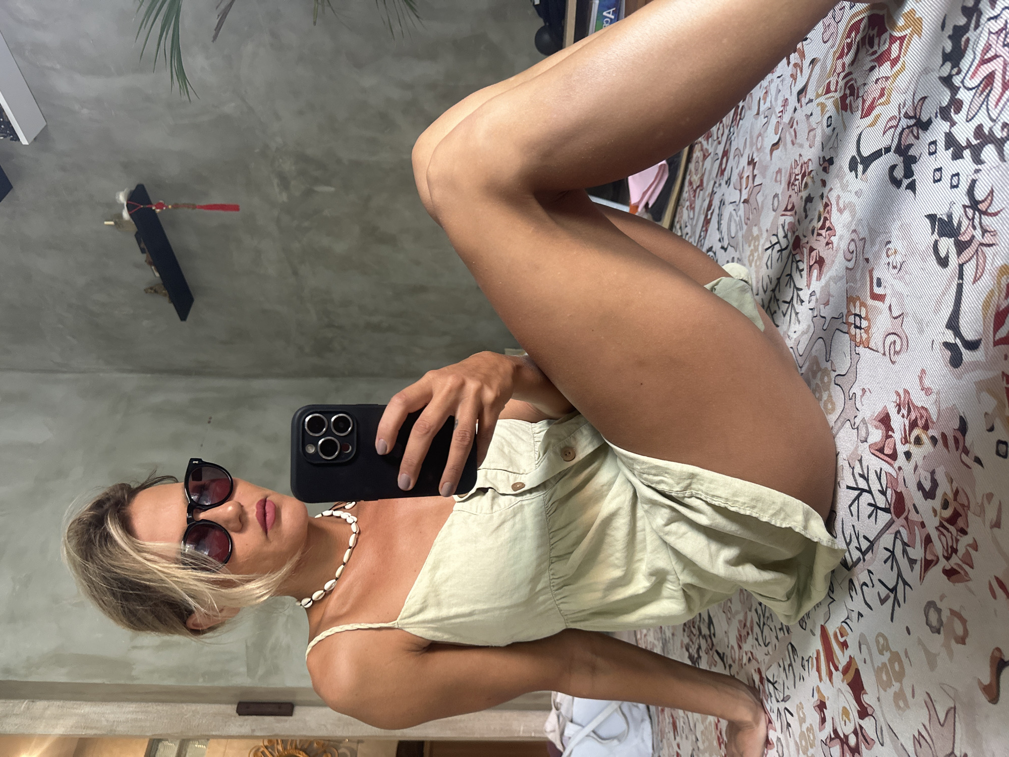 A woman wearing sunglasses, a white shell necklace, and a light green romper is taking a selfie with her phone in front of a mirror. She is lying on a bed with a patterned bedspread. The background shows a room with a concrete-like wall. In the upper left corner, there's a decorative object hanging on the wall. This photo was taken in North Kuta, Indonesia.