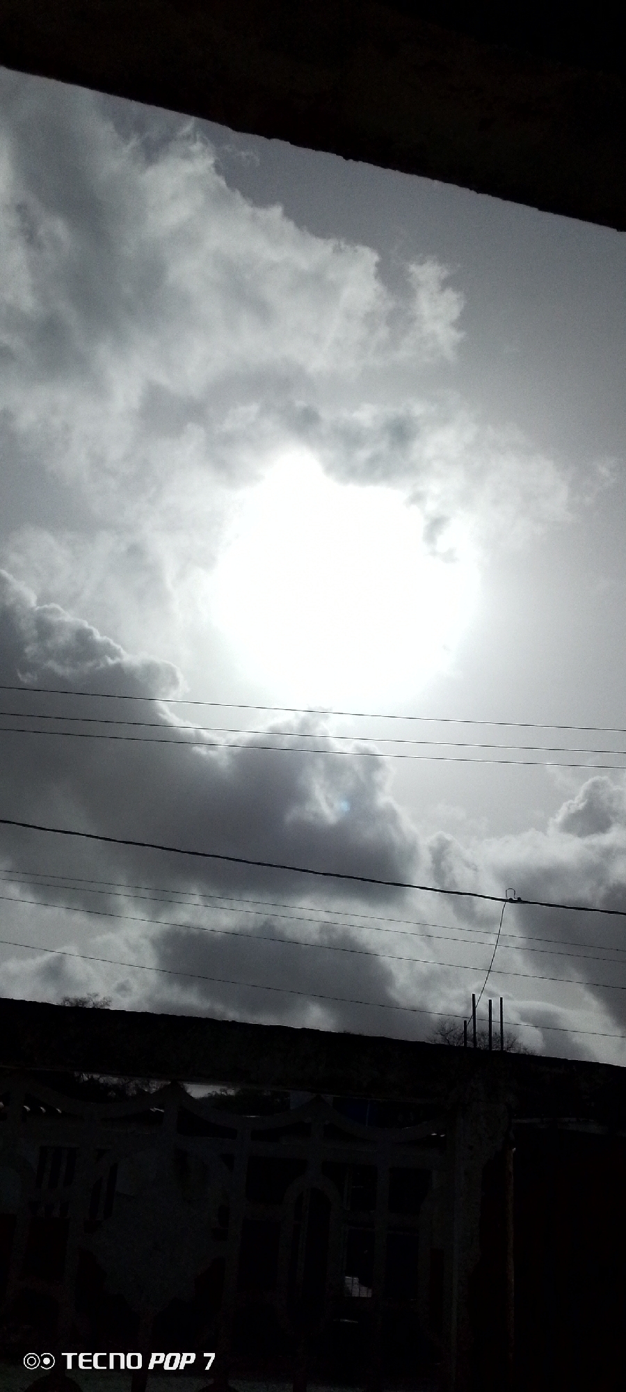 A black and white, low-angle shot of a cloudy sky with visible power lines. The bottom portion of the frame is obscured by a dark structure, possibly a building or wall with fencing. The word "TECNO POP 7" appears in the lower left corner. The location and time of day cannot be determined from the image.