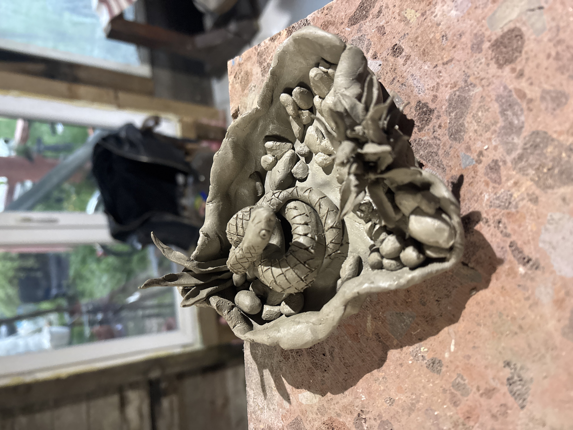 A high-angle, slightly out-of-focus, color photograph depicts a clay sculpture resting on a rough, reddish-brown surface with mottled coloration. The sculpture consists of a coiled clay snake positioned within a crude clay bowl-like structure. Small, rounded clay pieces, resembling pebbles, fill the space around the snake. The bowl has an uneven, roughly formed edge. To the left of the sculpture, a window is visible, showing a blurred outdoor scene with greenery and a white frame. Behind the sculpture and window, there are blurry objects, including what appears to be a black bag and a wooden structure. The lighting suggests an indoor setting with daylight. The image suggests the scene is in an indoor location in an unknown city; it does not provide information to confirm the location of Verinsko, Bulgaria.