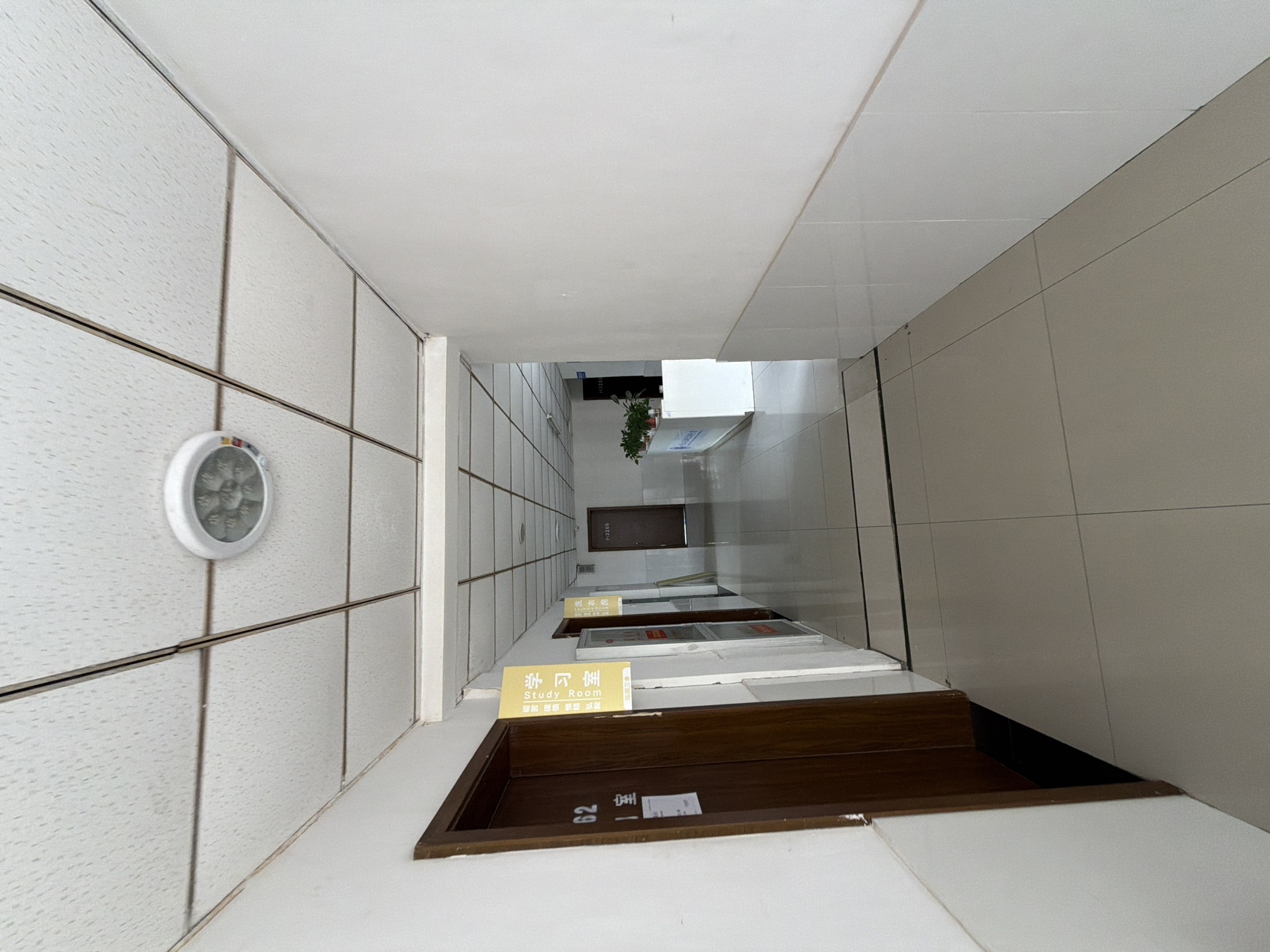 The image shows a view down a hallway in a building, possibly a school or office. The hallway has tiled floors and white walls. There are doors on either side of the hallway, some with signs. One sign, hanging from a yellow placard, reads "Study Room" in Chinese characters and English. Another door has the number "62" on it. Further down the hallway, a reception desk with plants is visible. The ceiling is a suspended tiled ceiling with a circular light fixture. The overall impression is of an indoor corridor in an institutional setting. The presence of Chinese text on signs suggests the location is in China. Given the context of the question, the image is likely from Jiangjin District, Chongqing, China.