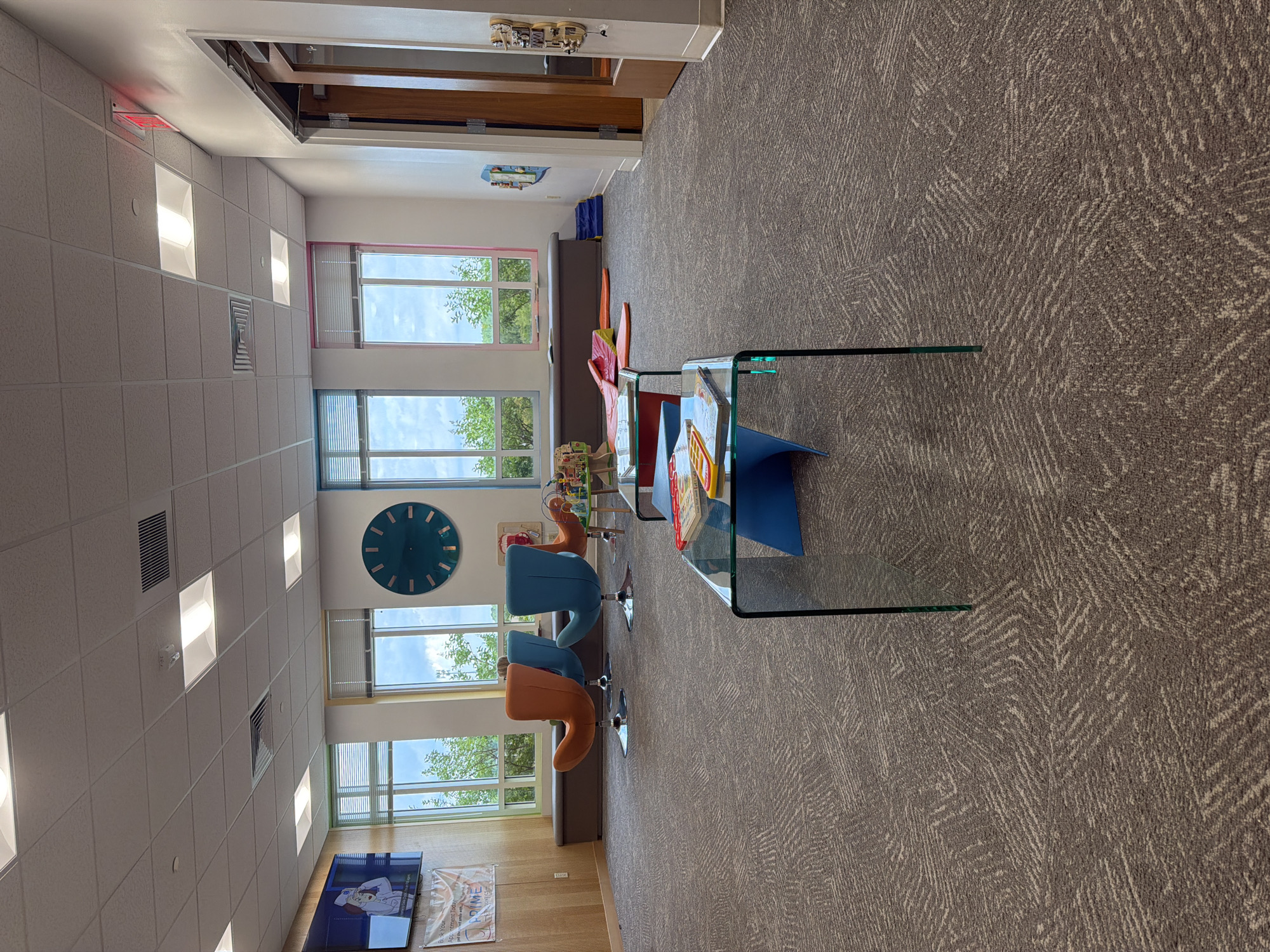 The image shows an interior view of what appears to be a waiting area or a children's play area. In the foreground, a low, glass-topped coffee table with a blue base holds several children's books. Beyond this, the room features a patterned carpet, several colorful, modern chairs, large windows with views of greenery, and a large, teal-colored clock on the wall. To the left, a flat-screen television displays a cartoon image. In the background, there are cushioned benches along the wall beneath the windows and some soft play mats on the floor. The overall atmosphere is bright and inviting, suggesting a public space designed for families. The specific location is the City of Chesapeake, United States of America.