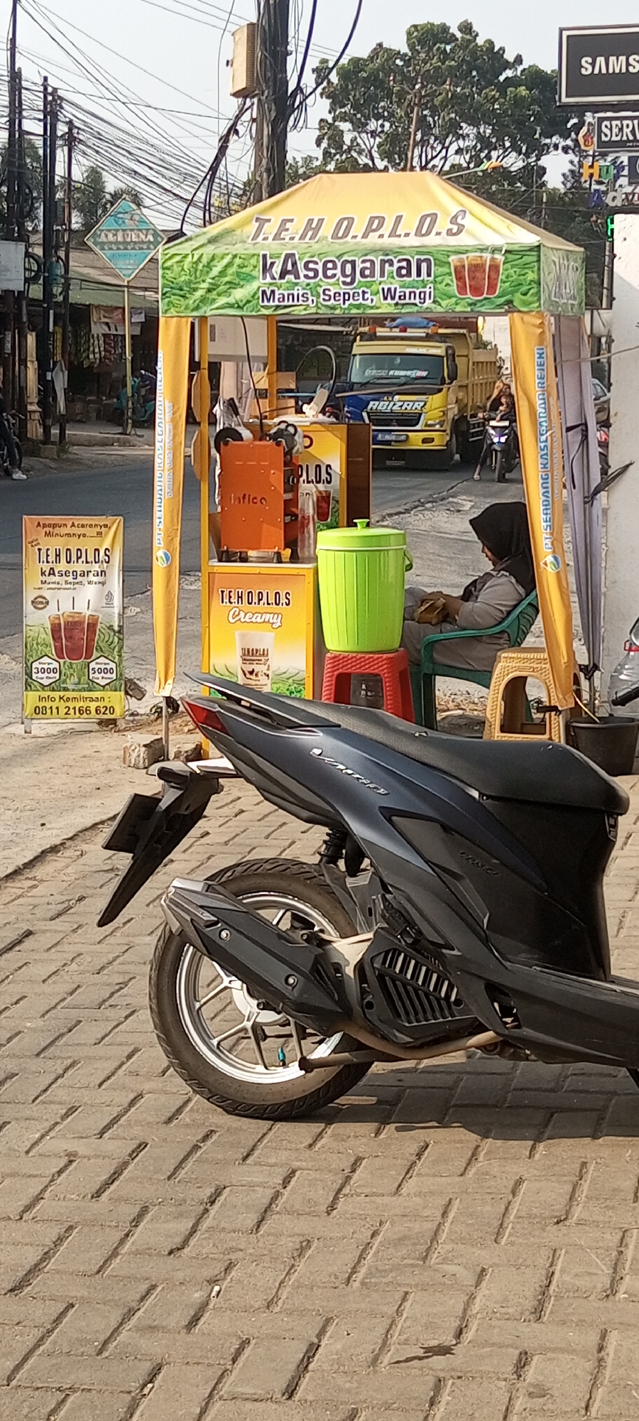 In the city of Setu, Indonesia, a roadside beverage stall operates under a yellow canopy advertising "T.E.H. O.P.L.O.S. kAsegbaran" which translates to "Refreshing Tea". The stall features an orange infusion machine and a bright green trash bin placed on a red stool. A woman wearing a black hijab and grey athletic wear is seated in a green plastic chair, holding a snack. A parked motorcycle with a dark grey body and silver wheels is positioned in the foreground, partially obscuring the stall's base and the paved ground. Behind the stall, a yellow truck with "ABIZAR" emblazoned on its side is visible, along with other elements of an urban street scene including utility poles, wires, and trees. To the left of the stall, a white sign with an image of three glasses of tea and pricing information (3000 and 5000) is displayed, along with a contact number for business inquiries. The overall atmosphere suggests a typical street-side business on a sunny day.