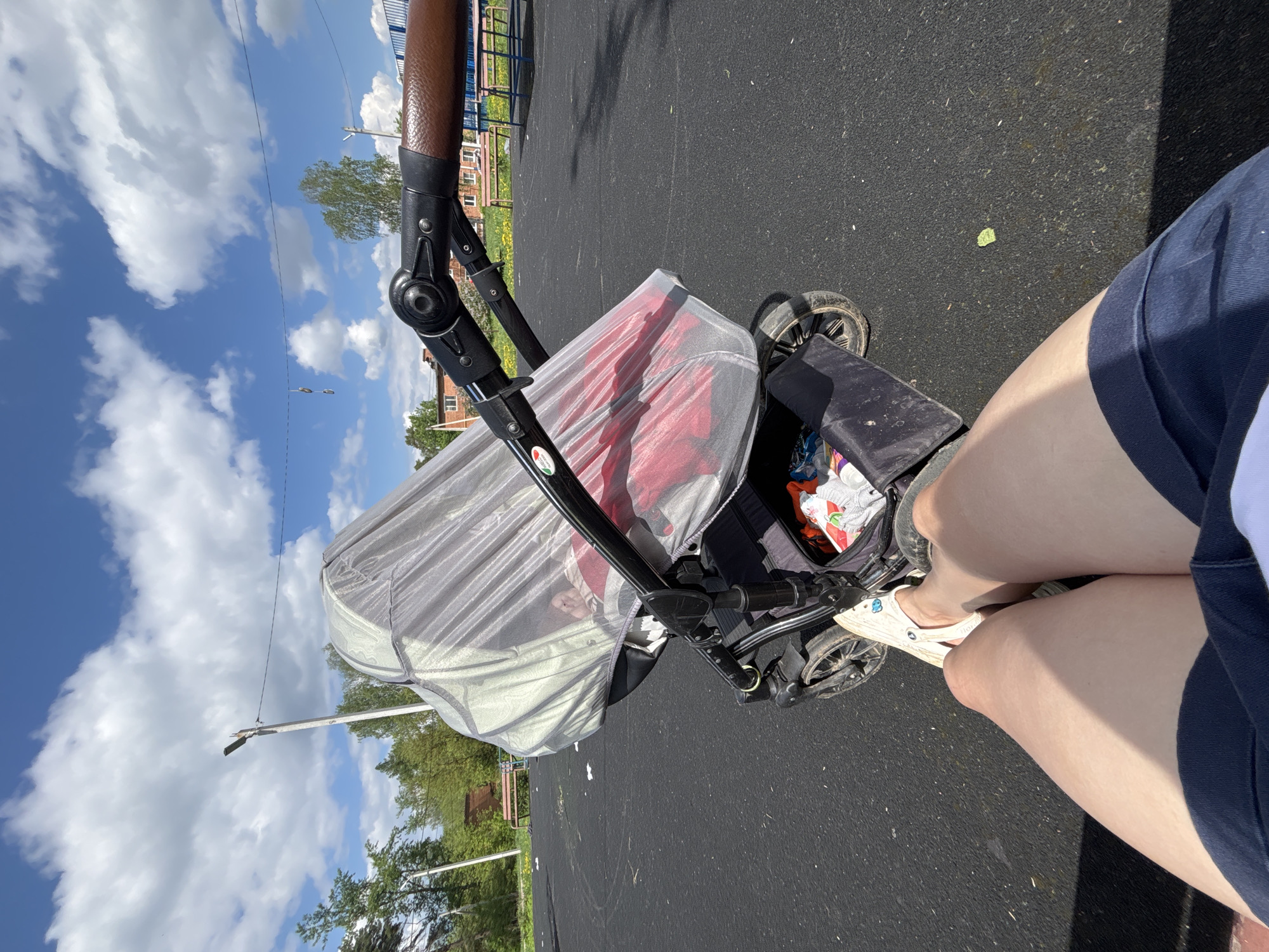 The image shows a first-person perspective, looking down at a stroller positioned on an asphalt surface. The stroller is covered with a mesh-like material and appears to contain a child. A person's legs and feet, wearing white Crocs, are visible next to the stroller. The background includes trees, buildings, and a blue sky with scattered clouds. A lamp post is also visible in the background. The image was taken outdoors, likely in Раздолье, Russia.