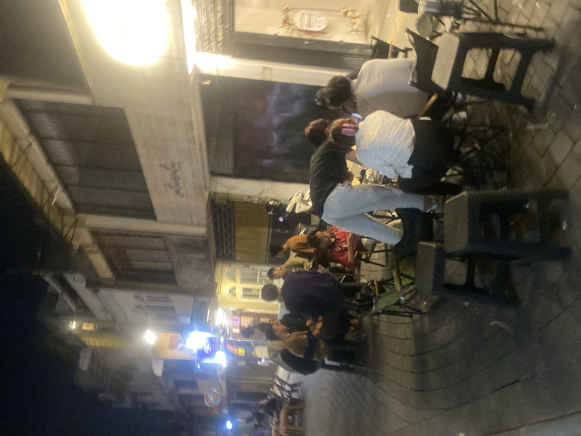 The image depicts an outdoor seating area at night, likely a cafe or restaurant, in Banjarmasin, Indonesia. The scene is illuminated by artificial lights, casting a warm glow on the patrons and the brick-paved ground. Several people are seated at small tables and chairs, engaged in conversation or looking at their phones. The architecture of the buildings in the background suggests a traditional urban setting, with multi-story structures and shops lining the street. A motorcycle is parked nearby, a common sight in Indonesian cities. The atmosphere appears relaxed and social, with people enjoying the evening outdoors.