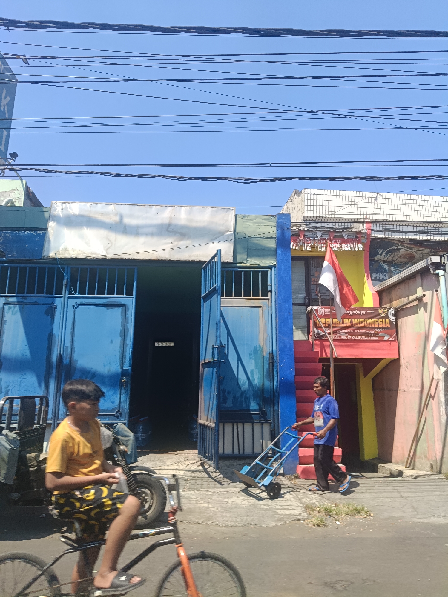 In Surabaya, Indonesia, a street scene unfolds under a clear blue sky, crisscrossed by numerous power lines. Dominating the foreground, a young boy, wearing a yellow t-shirt and patterned shorts, sits on a bicycle. To his right, a man in a blue t-shirt and dark pants pushes a blue hand truck along the sidewalk. Behind them, a building with a blue metal gate is visible, with an open doorway revealing a dark interior. A red staircase leads up to the second floor, where an Indonesian flag and banners are displayed, including one that reads "DIRGAHAYU REPUBLIK INDONESIA". The structures are a mix of industrial and residential in style, typical of urban Indonesian settings.