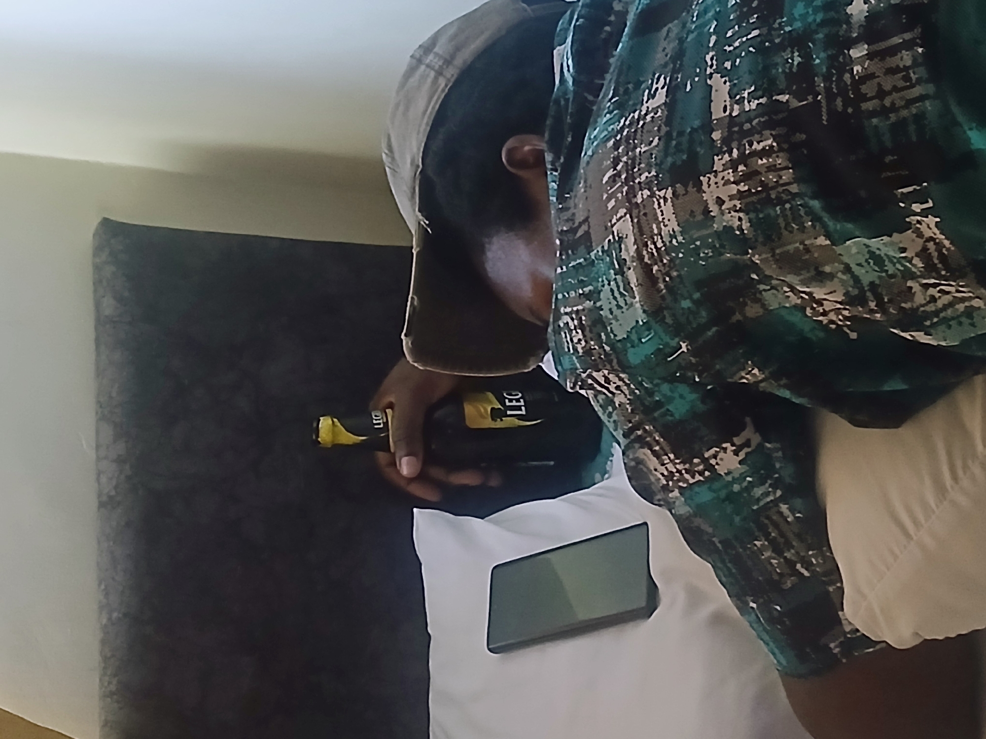 The scene depicts a man wearing a patterned shirt and a cap, holding a bottle of "Leg" beer. A smartphone lies on a white surface. The background includes a dark-colored wall. The city of Heipang, Nigeria, is not discernible from the image.