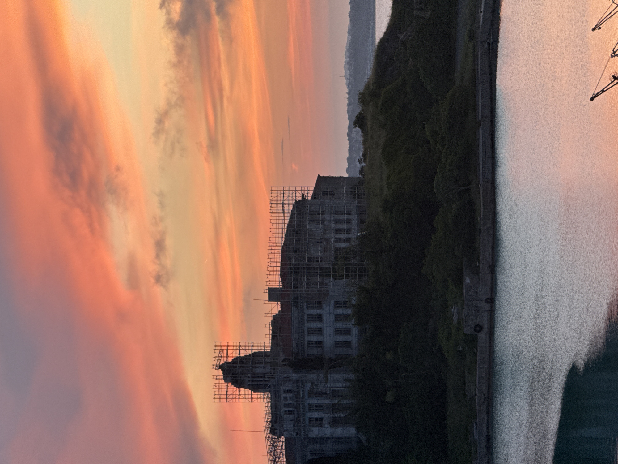 The image shows a dramatic sunset over a body of water, likely the sea, with a coastal town visible in the distance. The sky is ablaze with streaks of orange, pink, and yellow, indicating either the golden hour or a spectacular sunset. In the foreground, a large, old building, possibly a historical structure or a former industrial site, is shrouded in scaffolding, suggesting ongoing renovation or demolition. The building is silhouetted against the vibrant sky and is surrounded by dark, lush greenery on a hill.
To the right of the building and hill, a wide expanse of water reflects the colors of the sky, creating a shimmering, textured surface. The water appears calm, with subtle ripples. Along the edge of the water, a concrete embankment or pier is visible, with a road or pathway running alongside it. In the far right of the image, the dark shapes of industrial cranes can be seen, hinting at a port or harbor setting.
The overall mood is one of dramatic natural beauty juxtaposed with human intervention and industry. The time of day is clearly sunset, and the weather appears clear, allowing for the full spectacle of the sky's colors. The location context of Sozopol, Bulgaria, suggests a coastal European setting, known for its historical architecture and seaside beauty. There is no visible text in the image.