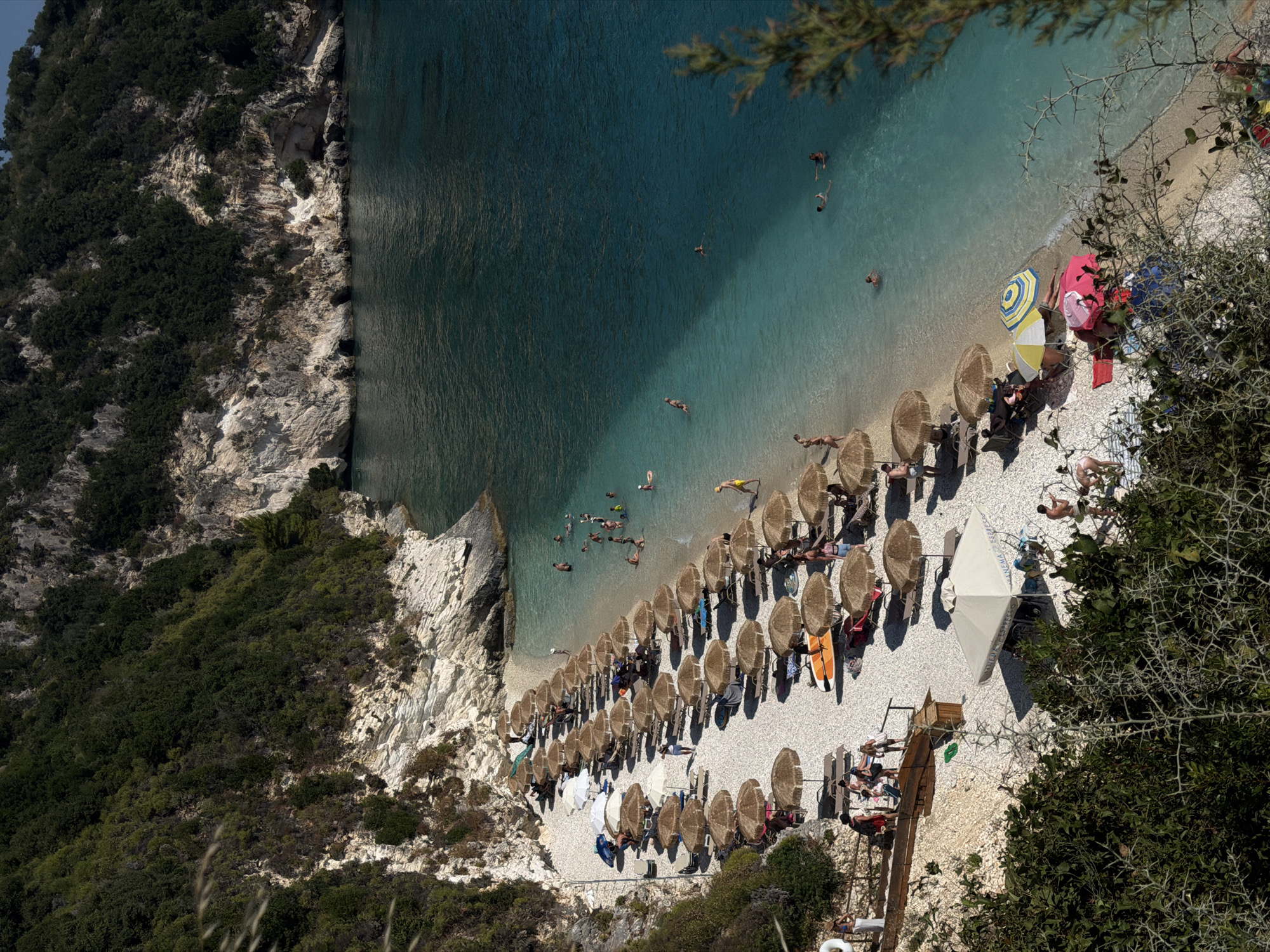 This is a high-angle, full-shot photograph of a crowded beach on a sunny day. The beach has white pebbles and is lined with rows of straw umbrellas and lounge chairs. The water is a clear turquoise, with several people swimming and playing in the shallow areas. In the background, lush green hills and rocky cliffs rise steeply from the shoreline.
There are numerous individuals on the beach, some relaxing under umbrellas, others sunbathing on lounge chairs, and some walking or playing near the water. A few people are seen swimming in the ocean. Beach accessories like towels, umbrellas, and lounge chairs are scattered across the pebble shore.
The environment is characterized by the natural beauty of a coastal landscape, with the stark white cliffs contrasting with the vibrant green vegetation and the brilliant blue of the sea. The overall scene depicts a popular summer destination with a lively atmosphere.