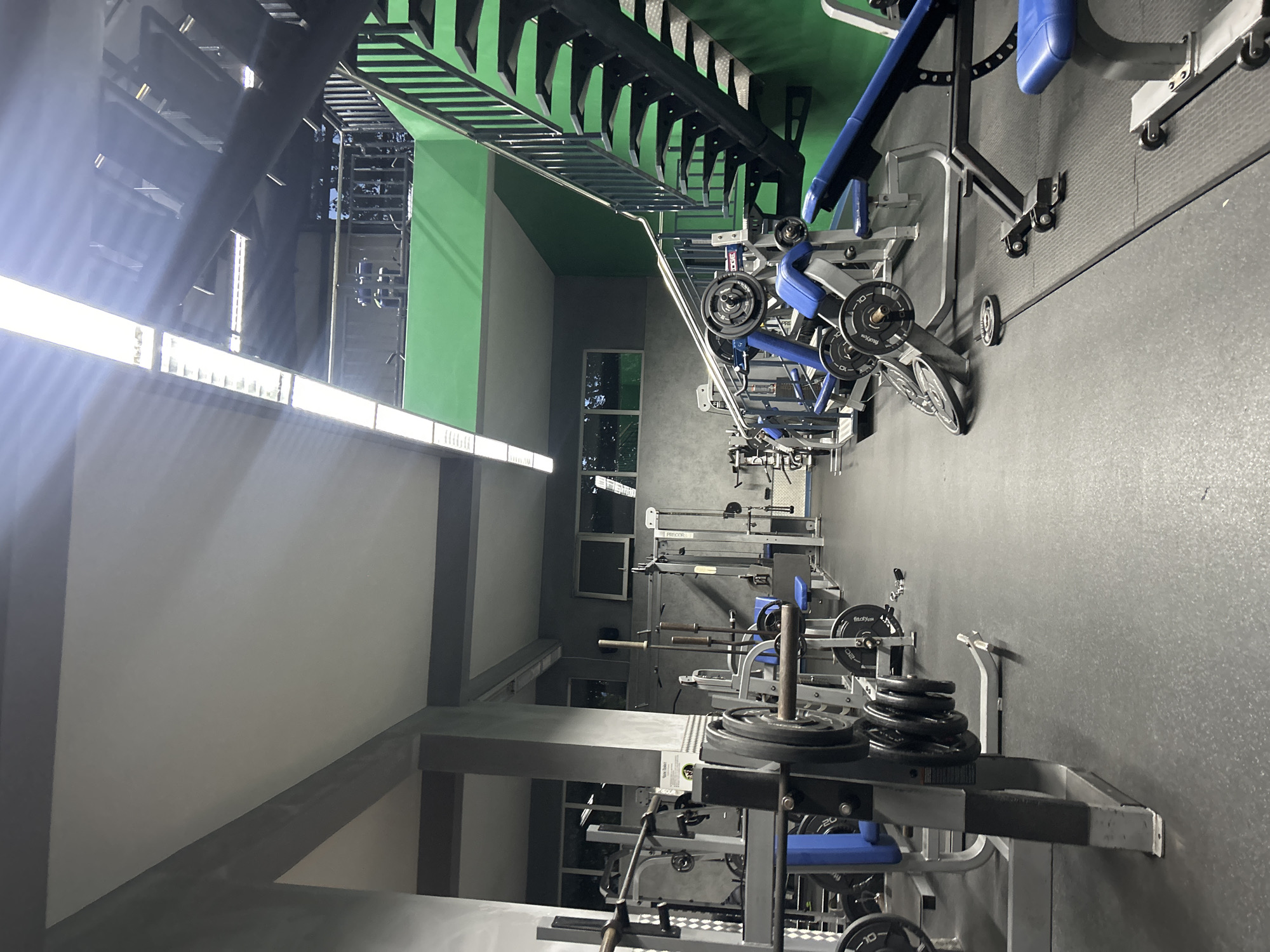 A gym in Burgas, Bulgaria. The interior features a variety of weightlifting machines and free weights arranged along a dark grey rubber floor. Equipment is predominantly blue, silver, and black. Overhead, exposed ductwork and lighting fixtures run along the ceiling. Natural light filters through windows. The gym appears unoccupied. The walls are painted in shades of green and grey. A metal staircase is visible on the side.