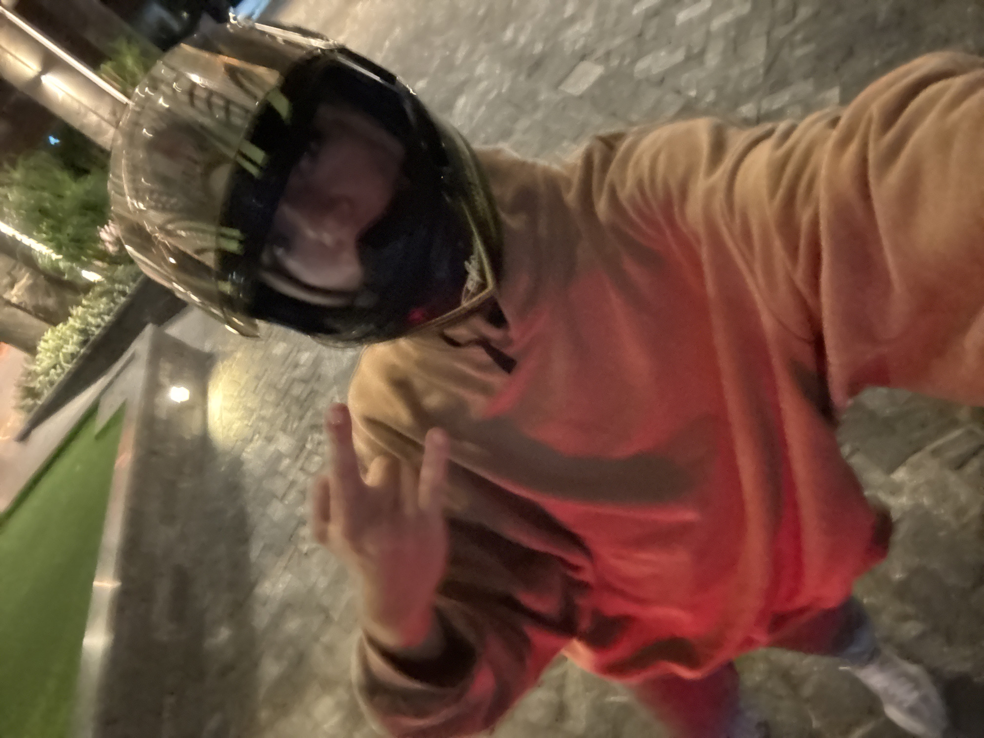 An individual wearing a black motorcycle helmet and an orange sweatshirt is posing outdoors, displaying a "peace" sign with their left hand. The background suggests a cityscape at night, with streetlights illuminating the surroundings. The ground is paved with what appears to be light-colored stone. The individual is wearing light-colored pants and shoes. Given the details visible, it's difficult to confirm the location definitively.